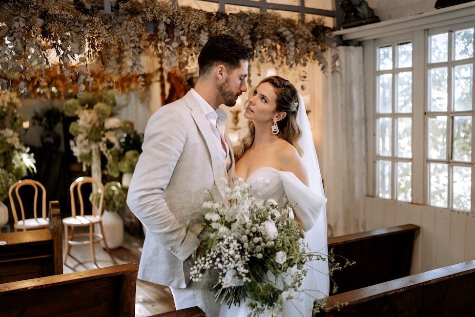 Ein Brautpaar bei einer Hochzeit, die Frau trägt ein weißes Brautkleid und hält einen Blumenstrauß, der Mann trägt einen hellen Anzug, beide schauen sich liebevoll an, im Hintergrund sind Blumen und Tische in einem festlichen Raum.
