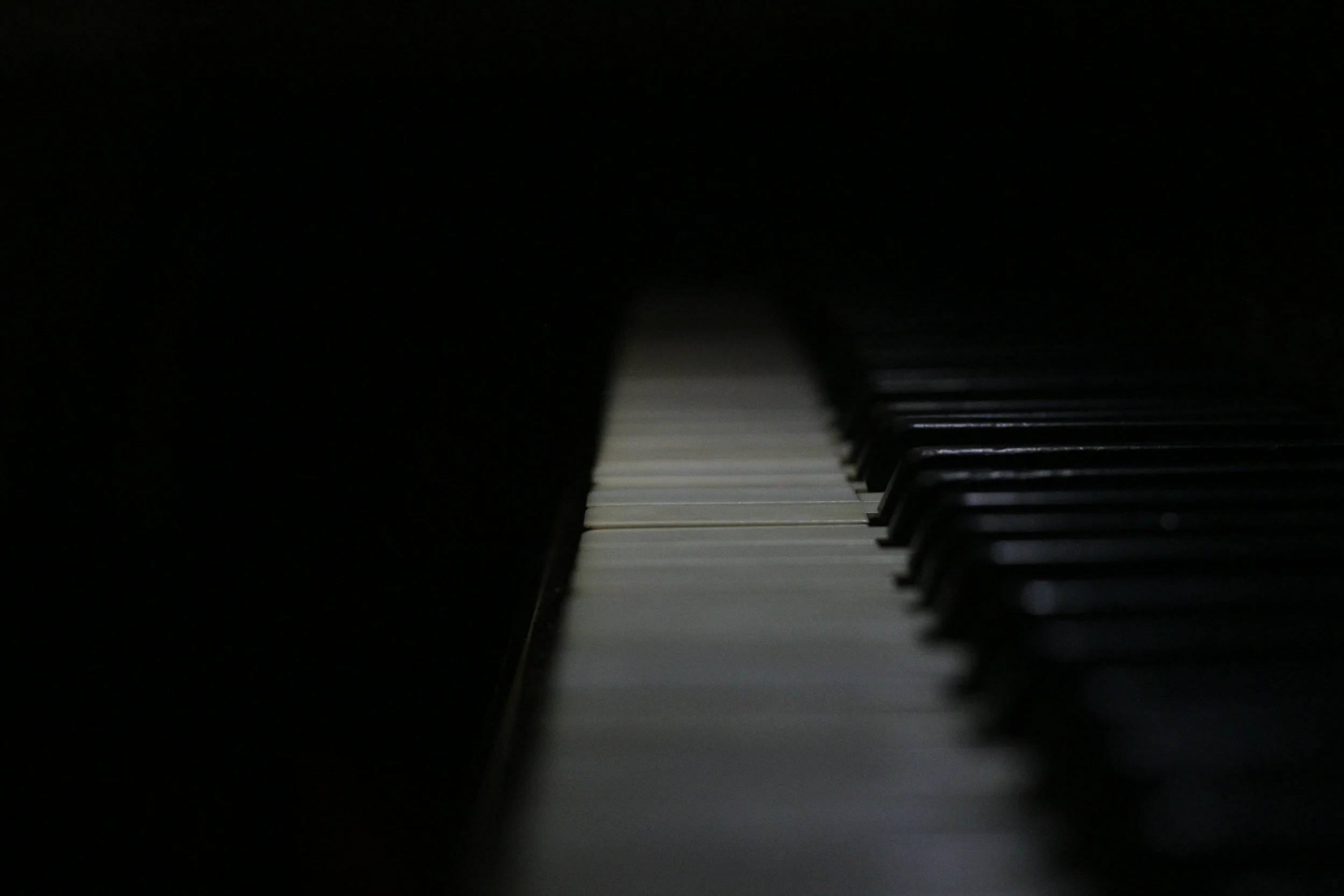 Close-up of piano keys, focusing on the white keys with black keys on the right, with soft lighting creating a shadowed effect.