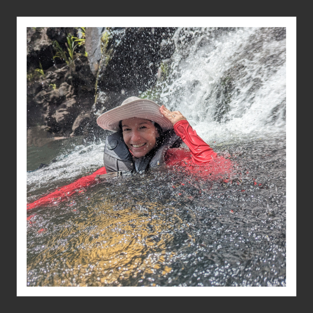 Instructor, Rachel, wearing a wide-brimmed hat and red waterproof jacket, smiling while swimming near a waterfall.