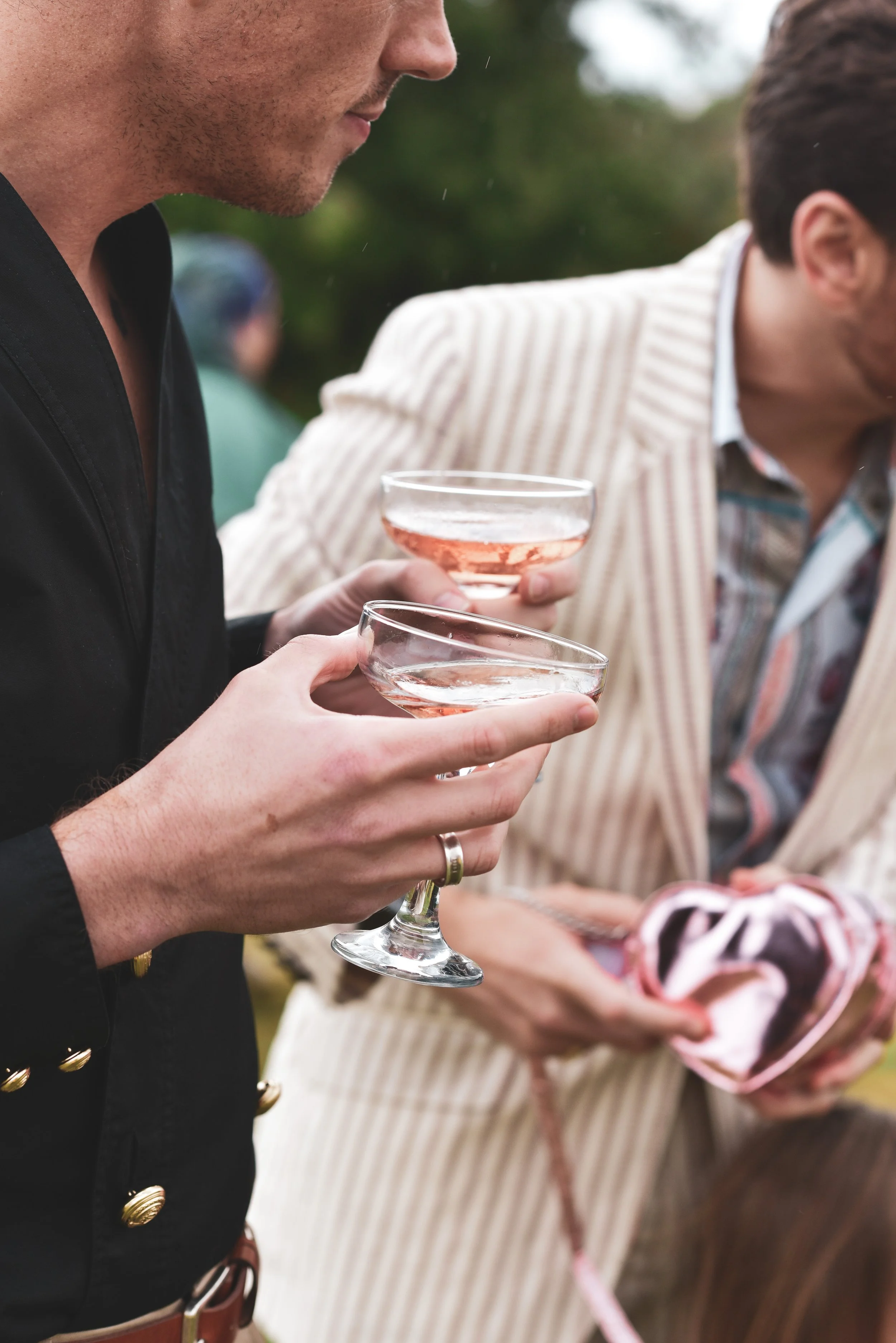 Close-up of people holding cocktail glasses at an outdoor gathering, with one person wearing a striped shirt and another in a dark blazer. The focus is on the hands and drinks, with blurred background.
