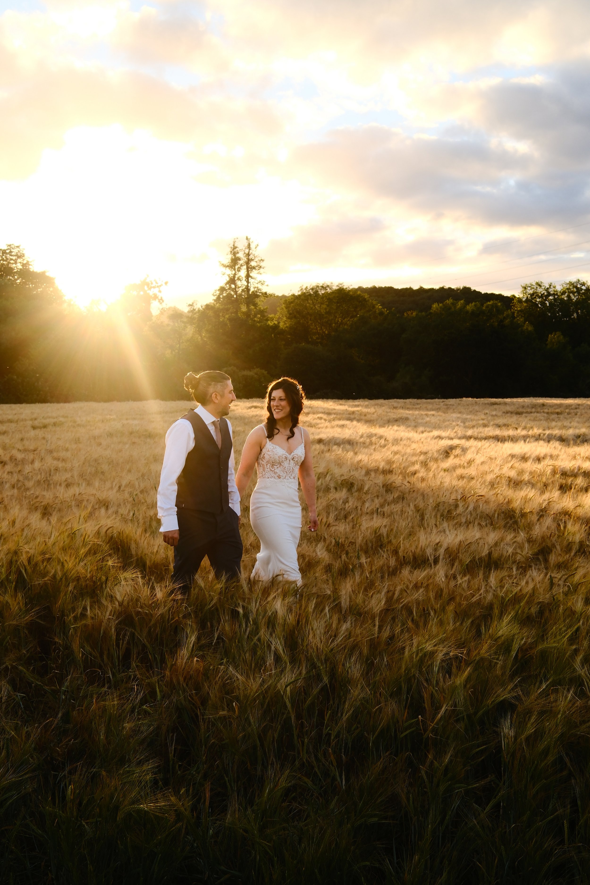 A couple dressed in wedding attire walking hand in hand through a field of tall grass during sunset, with trees and a cloudy sky in the background.