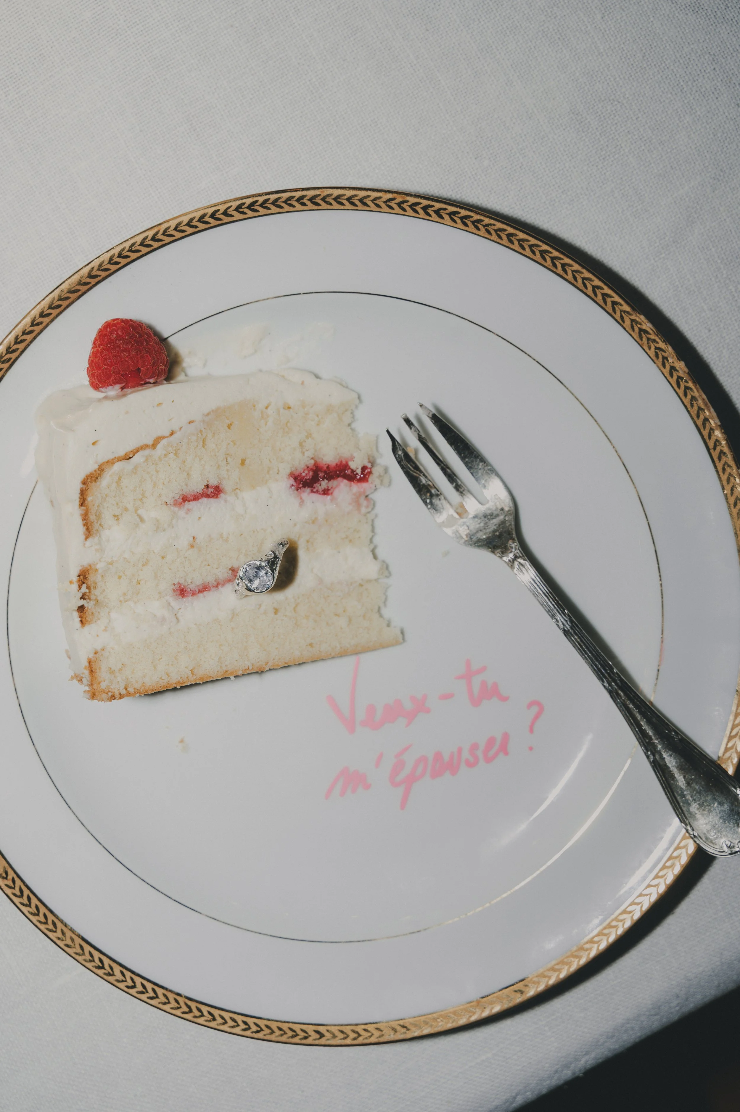 Une tranche de gâteau à la crème avec une framboise dessus et un petit bijou en forme de cœur sur la gâteau, une fourchette à côté, et la phrase 'Voues-tu m'épouser?' écrite en rose sur l'assiette.