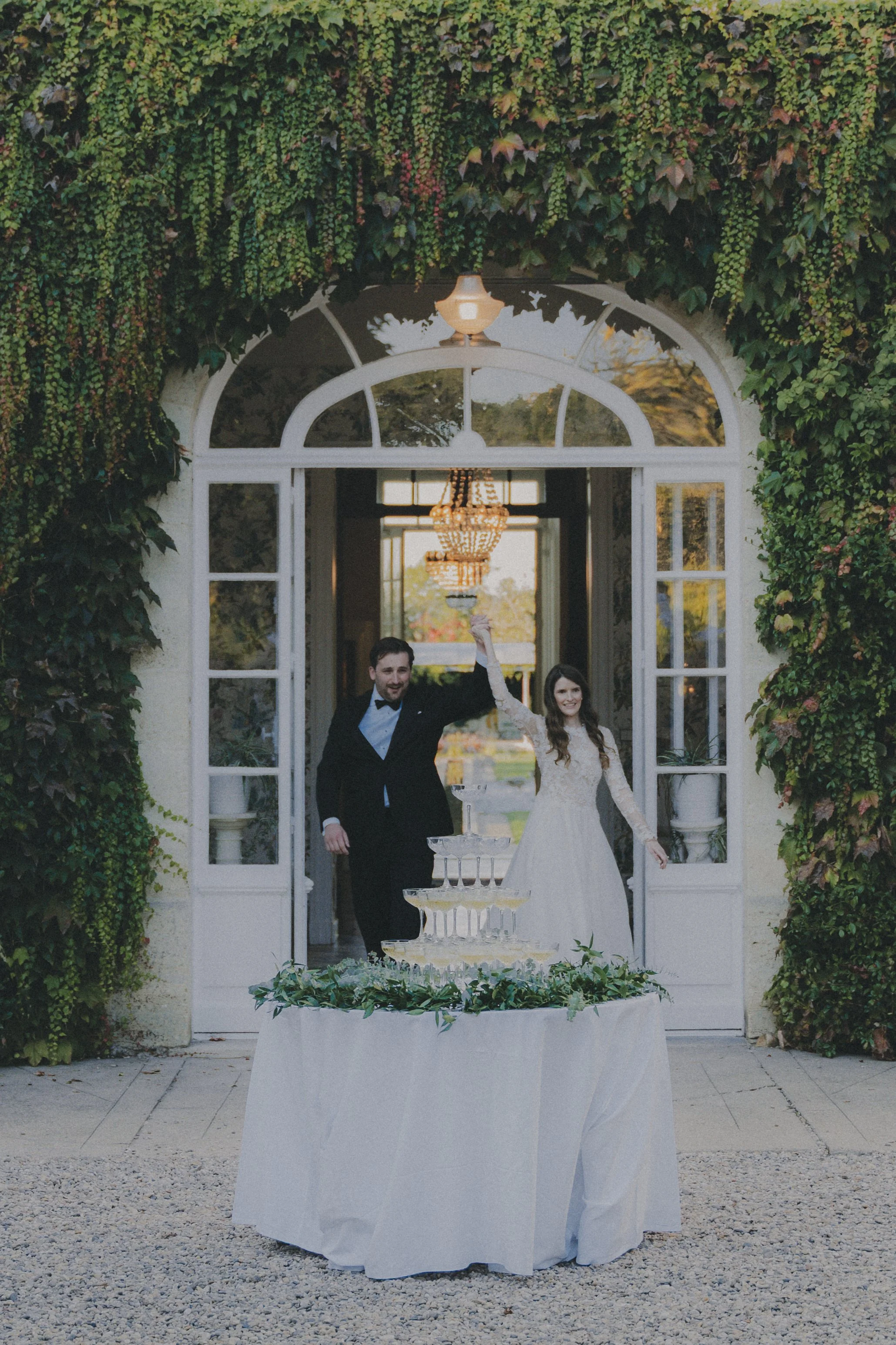 Un couple de mariés souriants sortant d'une bâtisse ornée de végétation, célébrant leur mariage avec un toast au champagne devant une pyramide de flûtes à champagne.