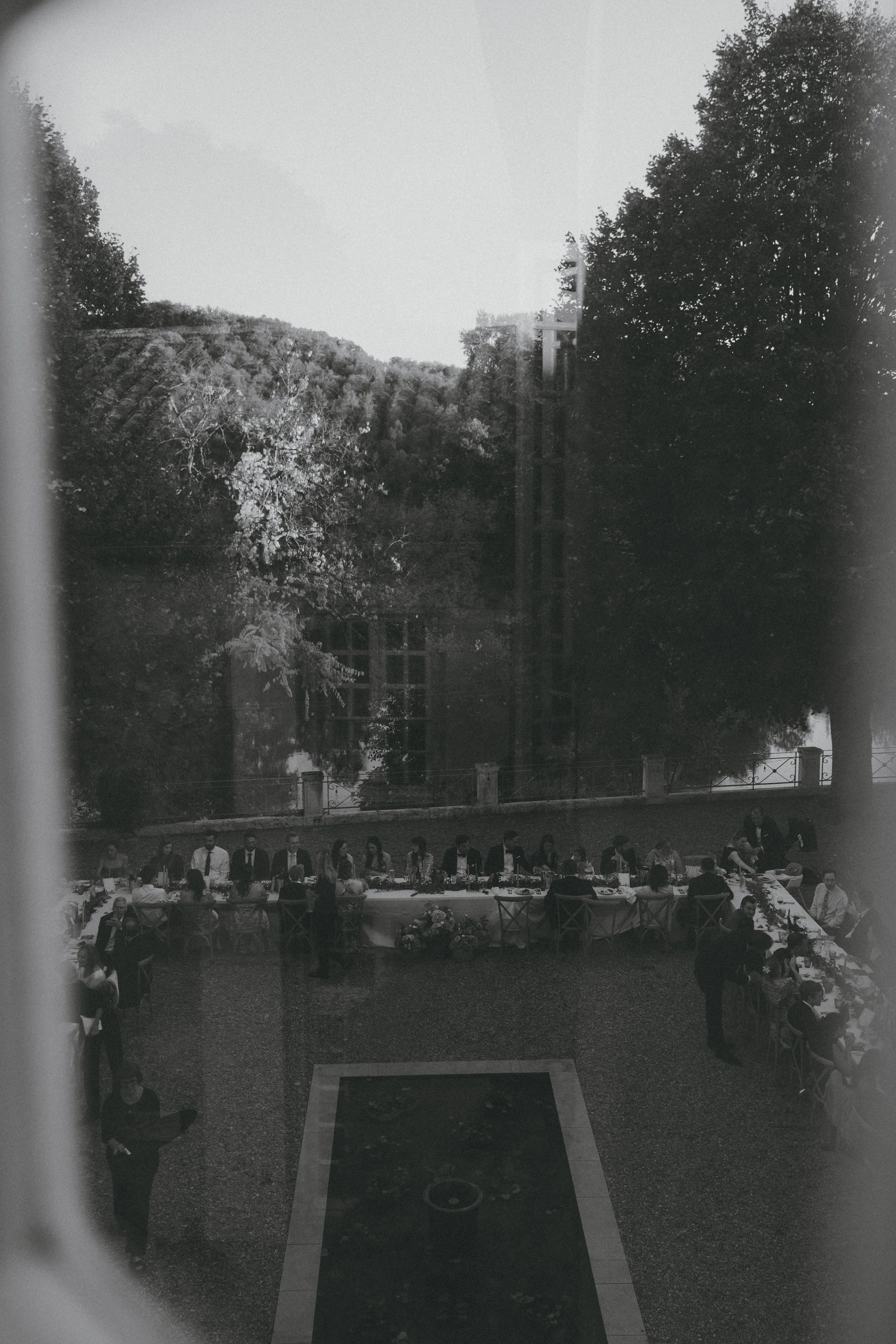 Une scène en noir et blanc montrant un rassemblement ou un dîner en plein air avec une longue table où des personnes sont assises, entourée d'arbres et d'une barrière en arrière-plan. Il y a également des personnes debout près de la table.