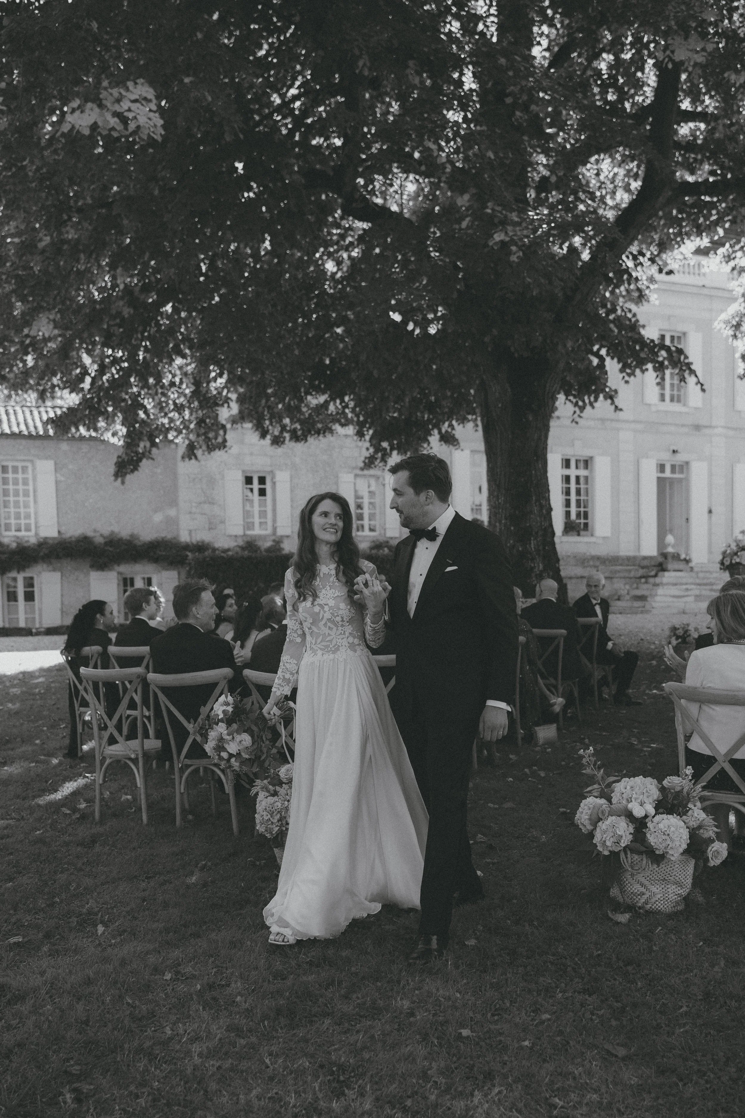 Un mariage en plein air avec un couple en tenues de mariage, la mariée en robe blanche et le marié en costume noir, échangeant un regard sous un arbre, entourés d'invités assis à l'extérieur.
