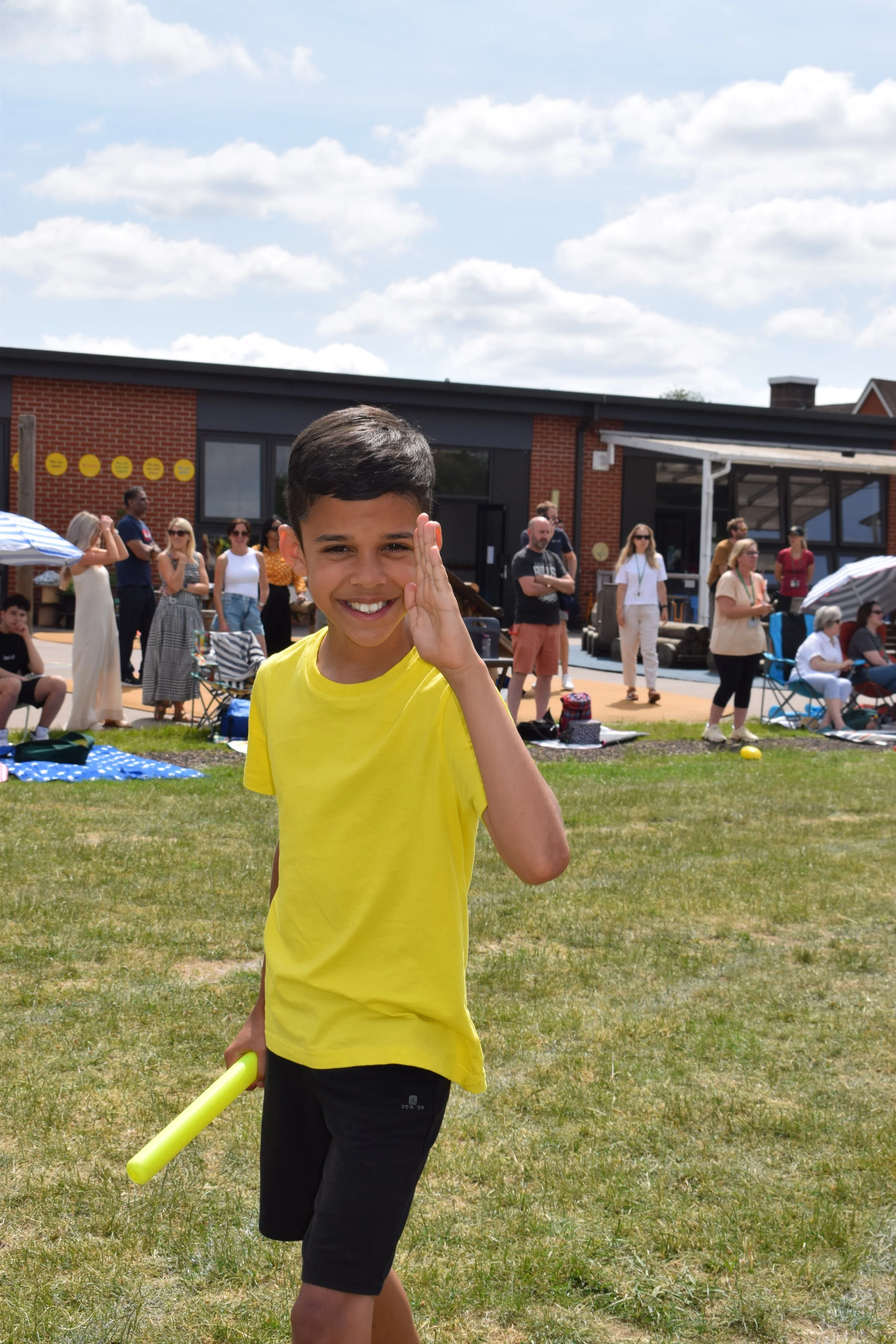 A young boy in a yellow shirt and black shorts holding a yellow stick, smiling and waving at the camera outdoors during a sunny day. People are gathered in the background, some sitting and some standing, near a building with a brick and black exterior, with umbrellas and lawn chairs on the grass.