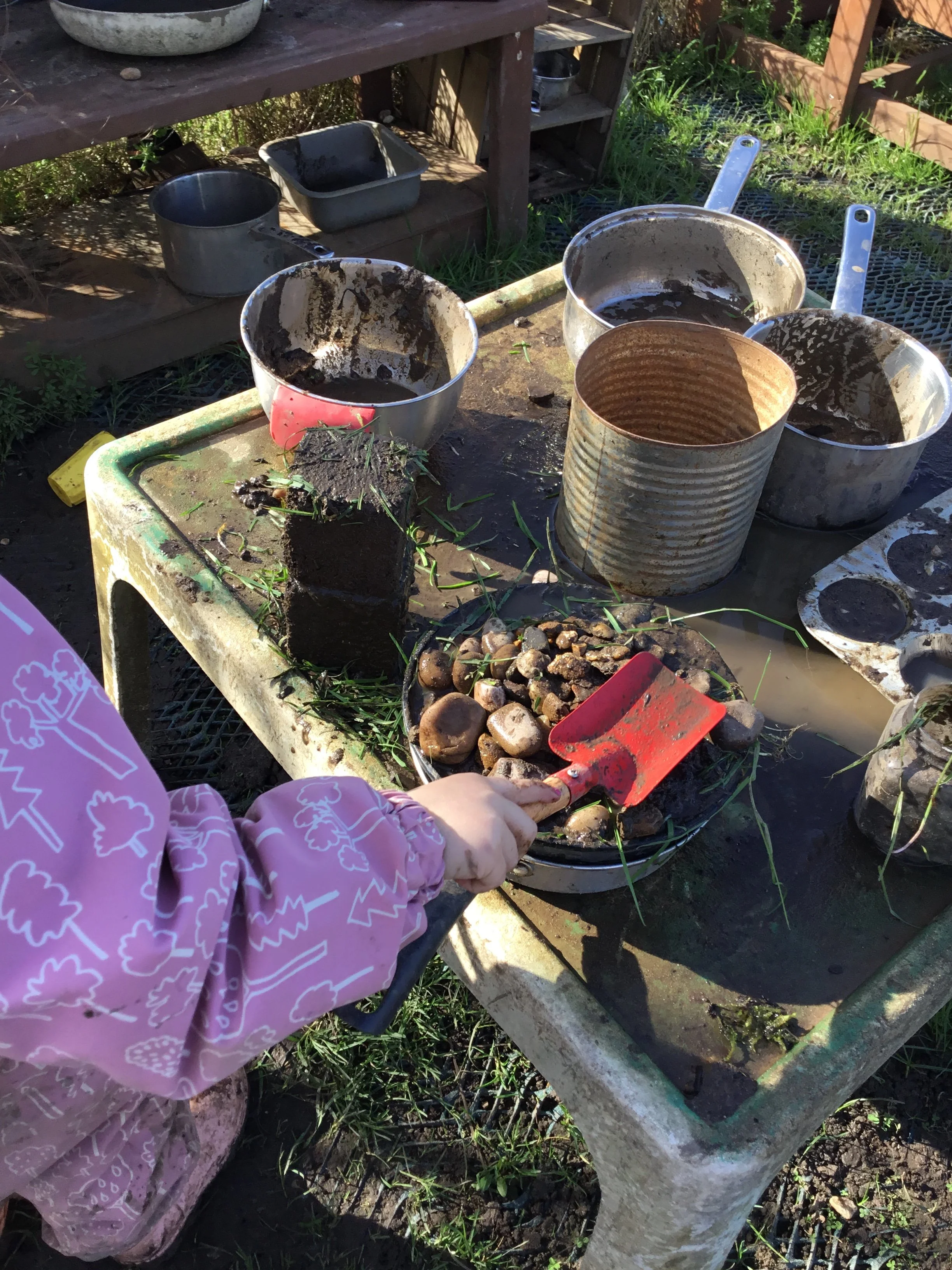 A person wearing a pink jacket with white patterns is using a small garden shovel to transfer soil or compost into a black container. The scene takes place outdoors on a worktable with various gardening tools, containers, and stones. Shelving units and greenery are visible in the background.