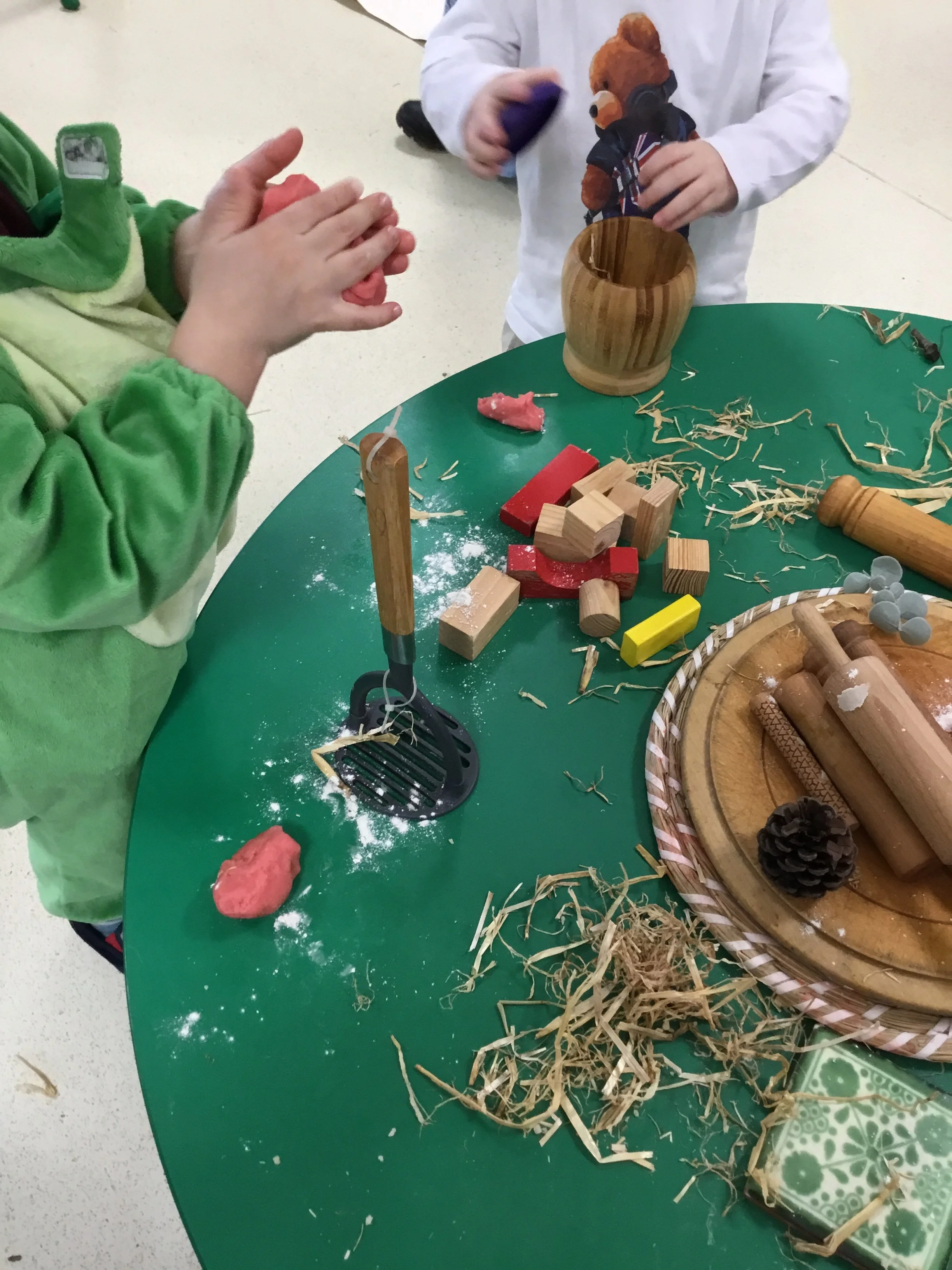 A child's hands clapping, with their face not visible. They are wearing a green jacket with yellow and cream accents. On the green table, there are scattered wooden blocks, shredded paper or straw, a red modeling clay, and a wooden rolling pin. Another child's torso and arms are visible, wearing a white long-sleeve shirt and holding a purple object.