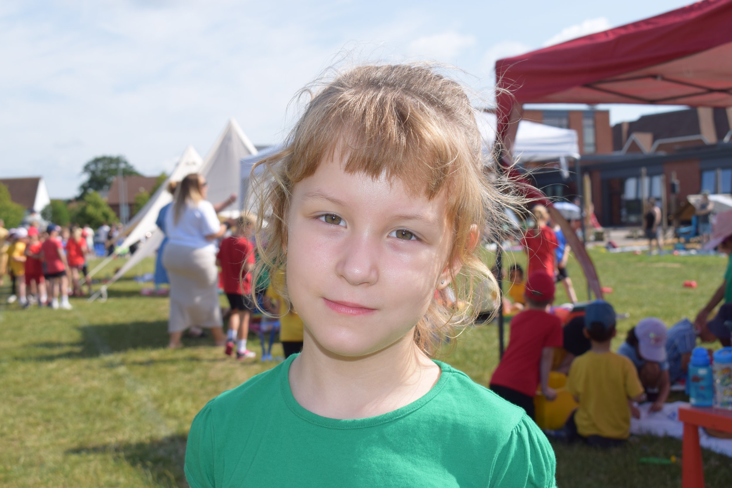 A young girl with blond hair and green eyes standing outdoors at a fair or festival, with tents and a crowd of children in the background.