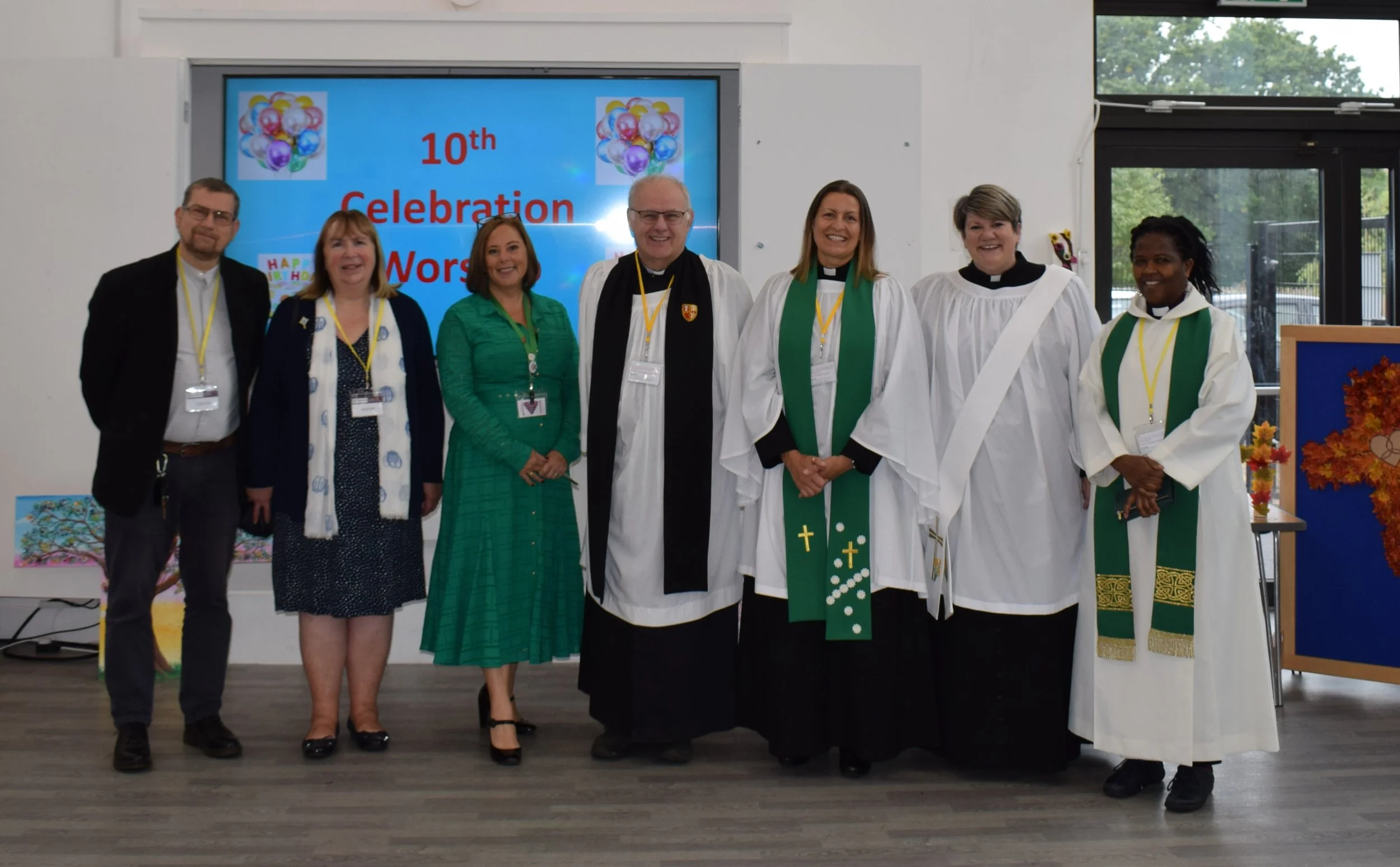 Group of seven people standing in front of a digital screen displaying a message about a 10th celebration anniversary. Some are wearing church vestments, and everyone is smiling.