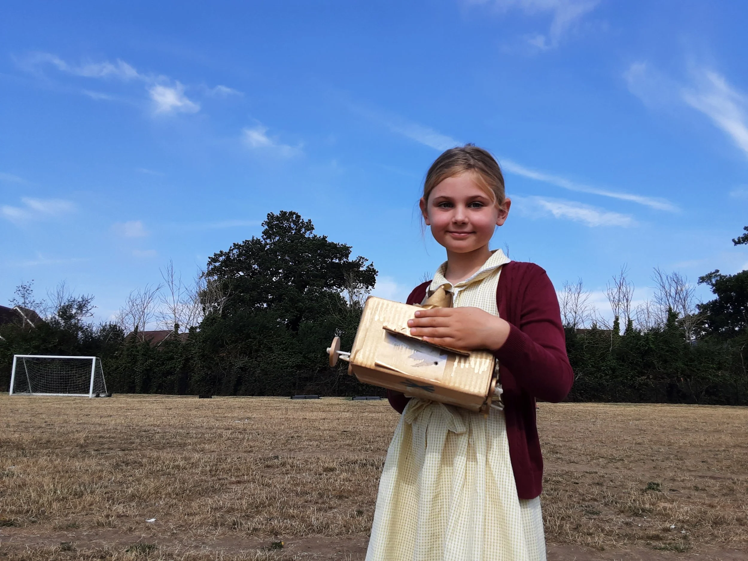 A young girl with blonde hair in a ponytail wearing a yellow checkered dress and maroon cardigan holding a cardboard box with cupcakes, standing on a soccer field with goalposts and trees in the background.