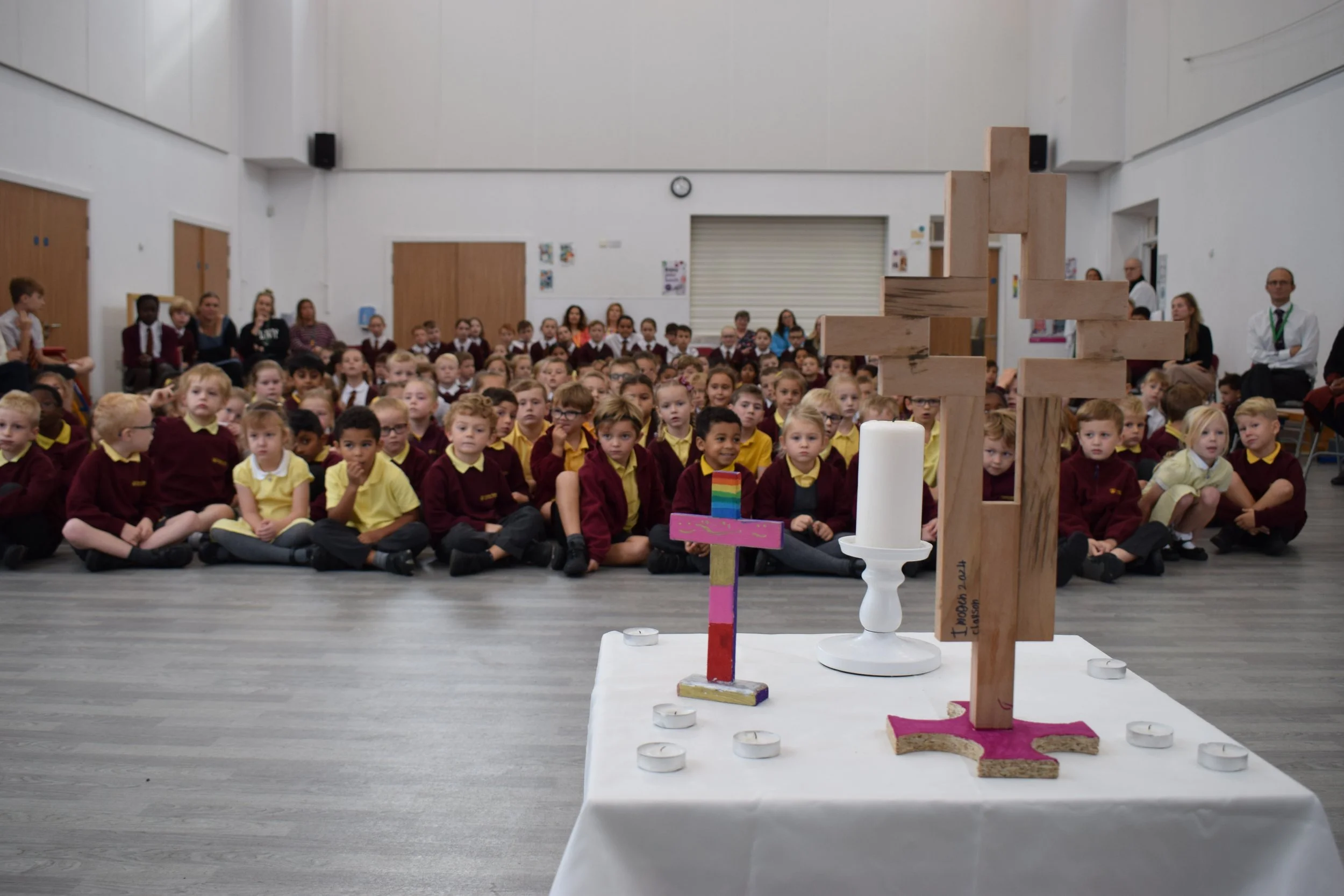 Children seated on the floor watching a structure built from wooden blocks on a table in a classroom or assembly hall.