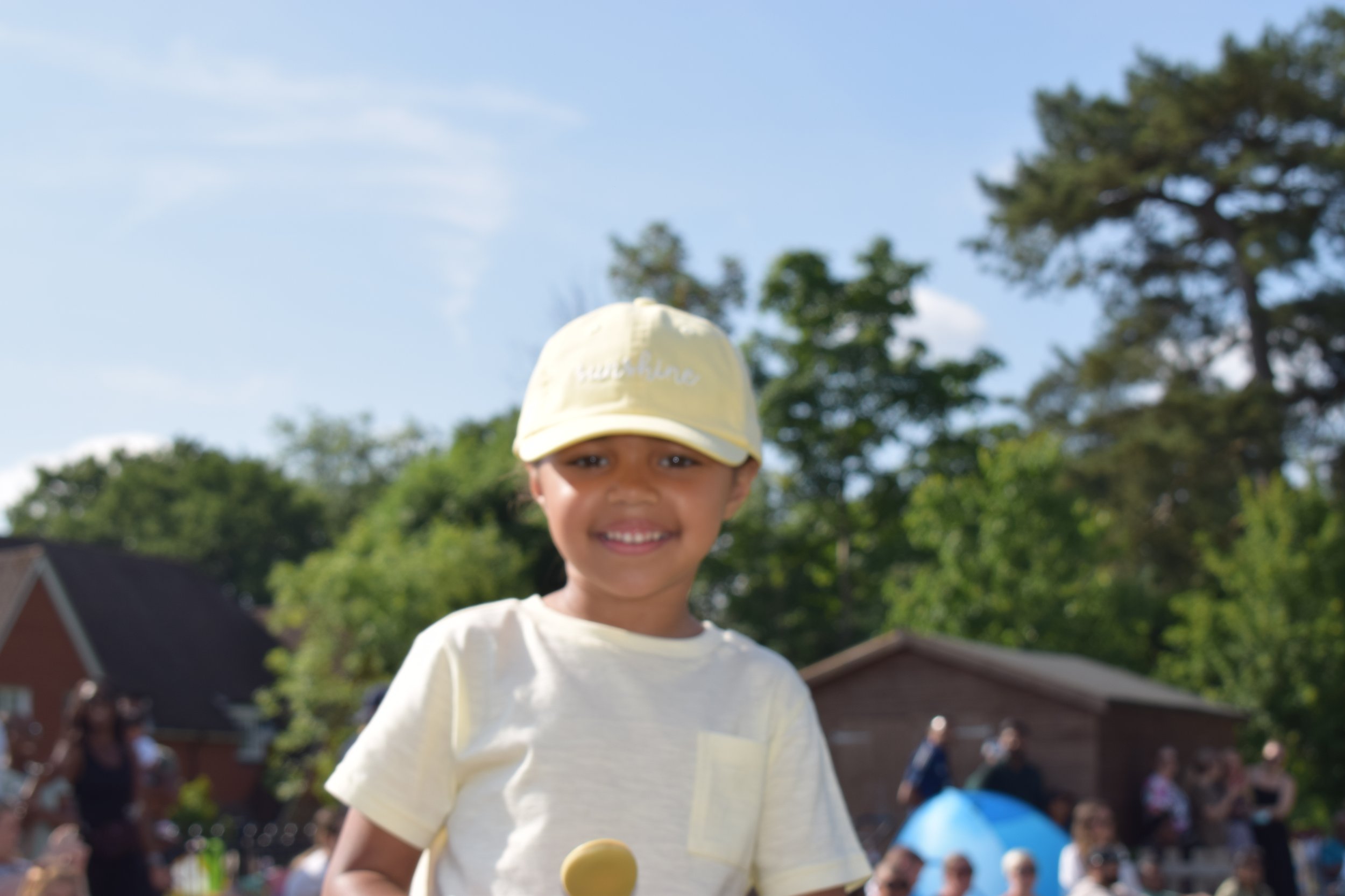 Young boy smiling outdoors during a sunny day, wearing a yellow hat and a white T-shirt, with trees and a crowd of people in the background.