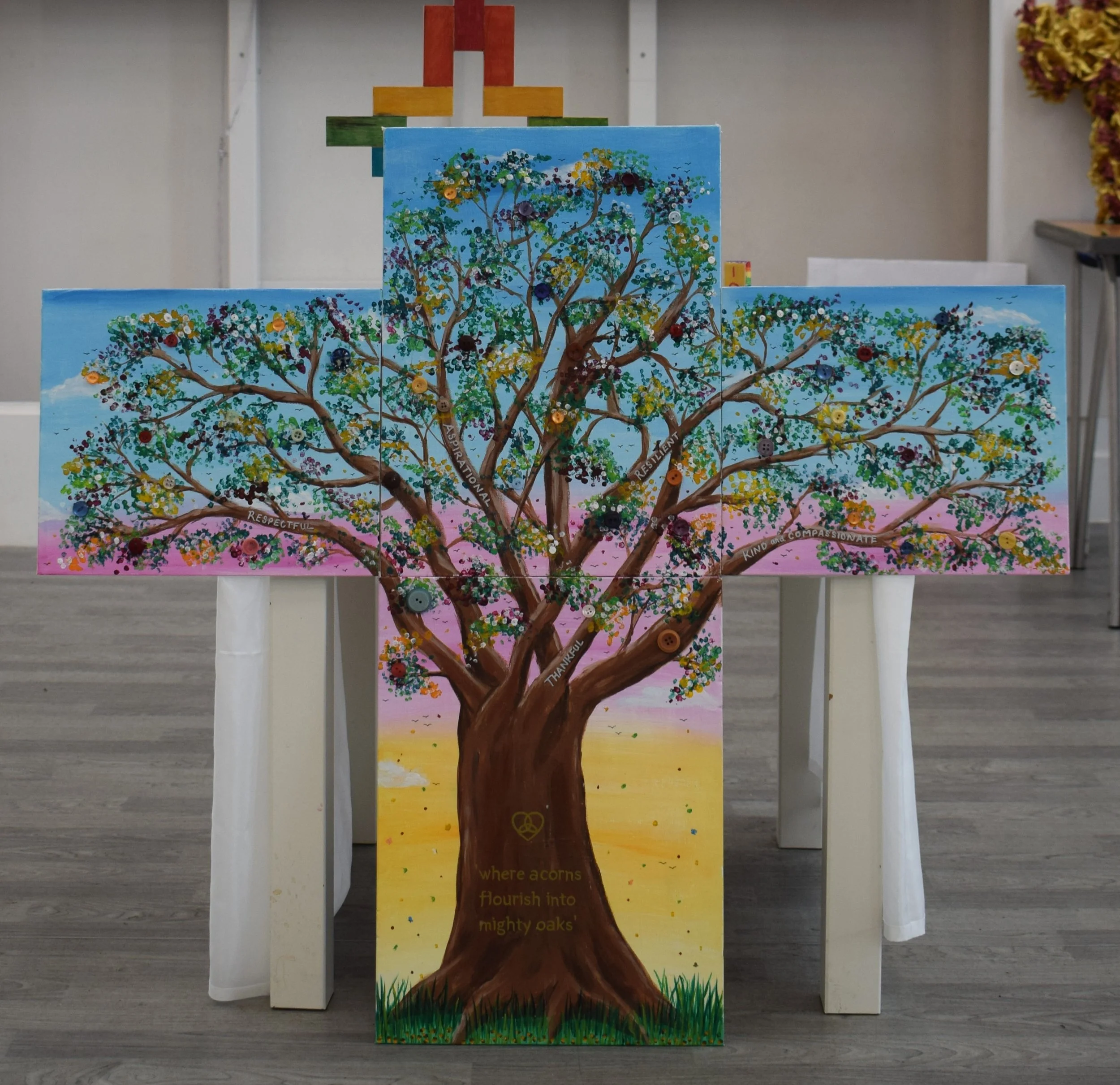 A colorful painted wooden artwork of a large tree with numerous green, yellow, purple, and white leaves set against a blue sky background. The tree has a brown trunk with words written on it, including "RESPECTFUL,"