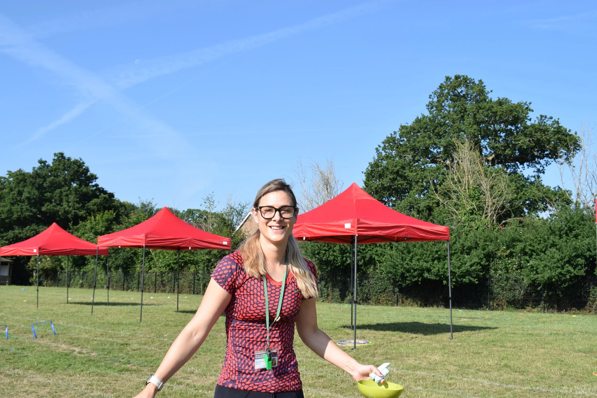 A woman with glasses and long blonde hair smiling outdoors under a clear blue sky, standing in front of red tents with trees in the background, holding a small yellow bowl and some papers.