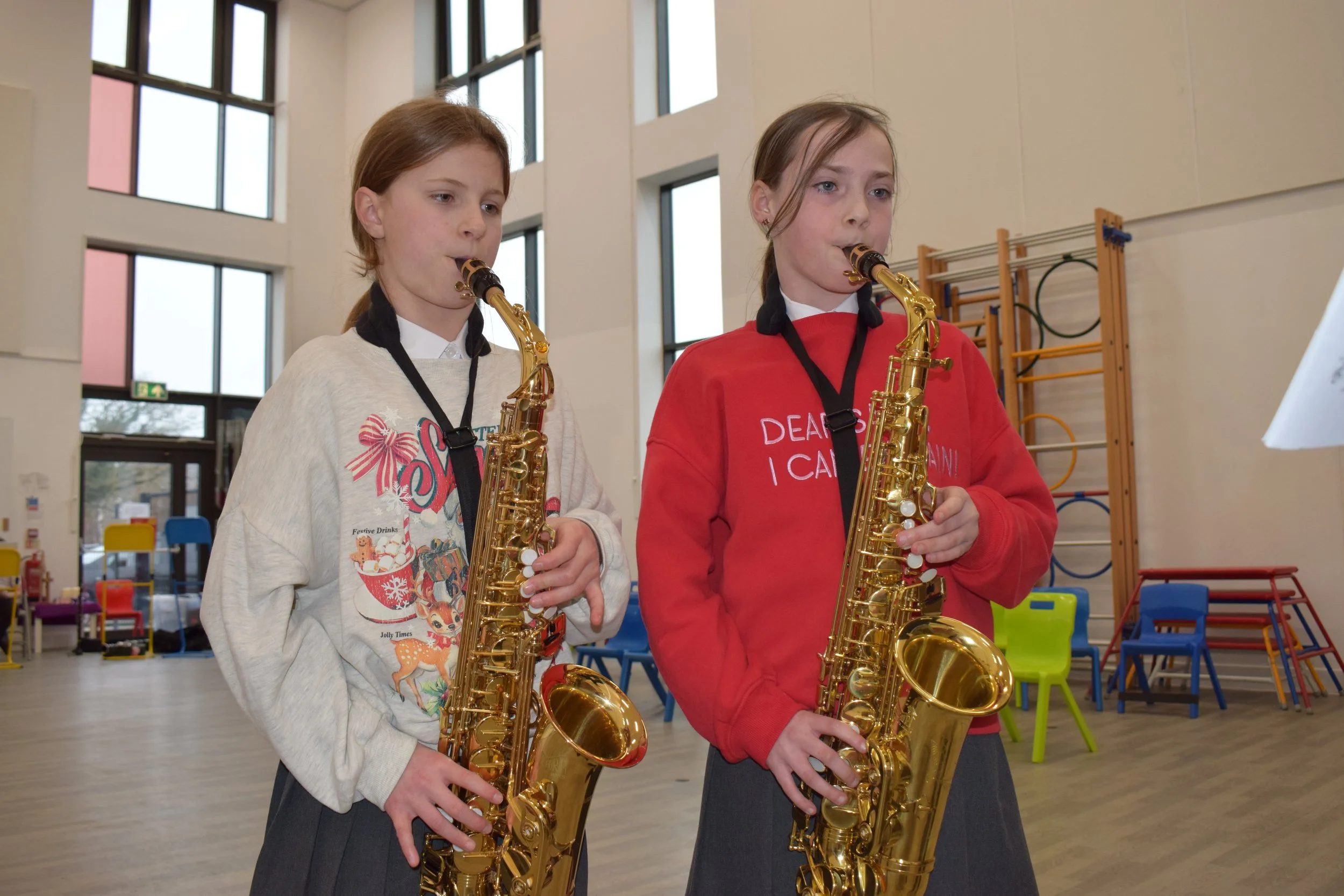 Two young girls playing saxophones in a spacious indoor room with large windows and colorful chairs.