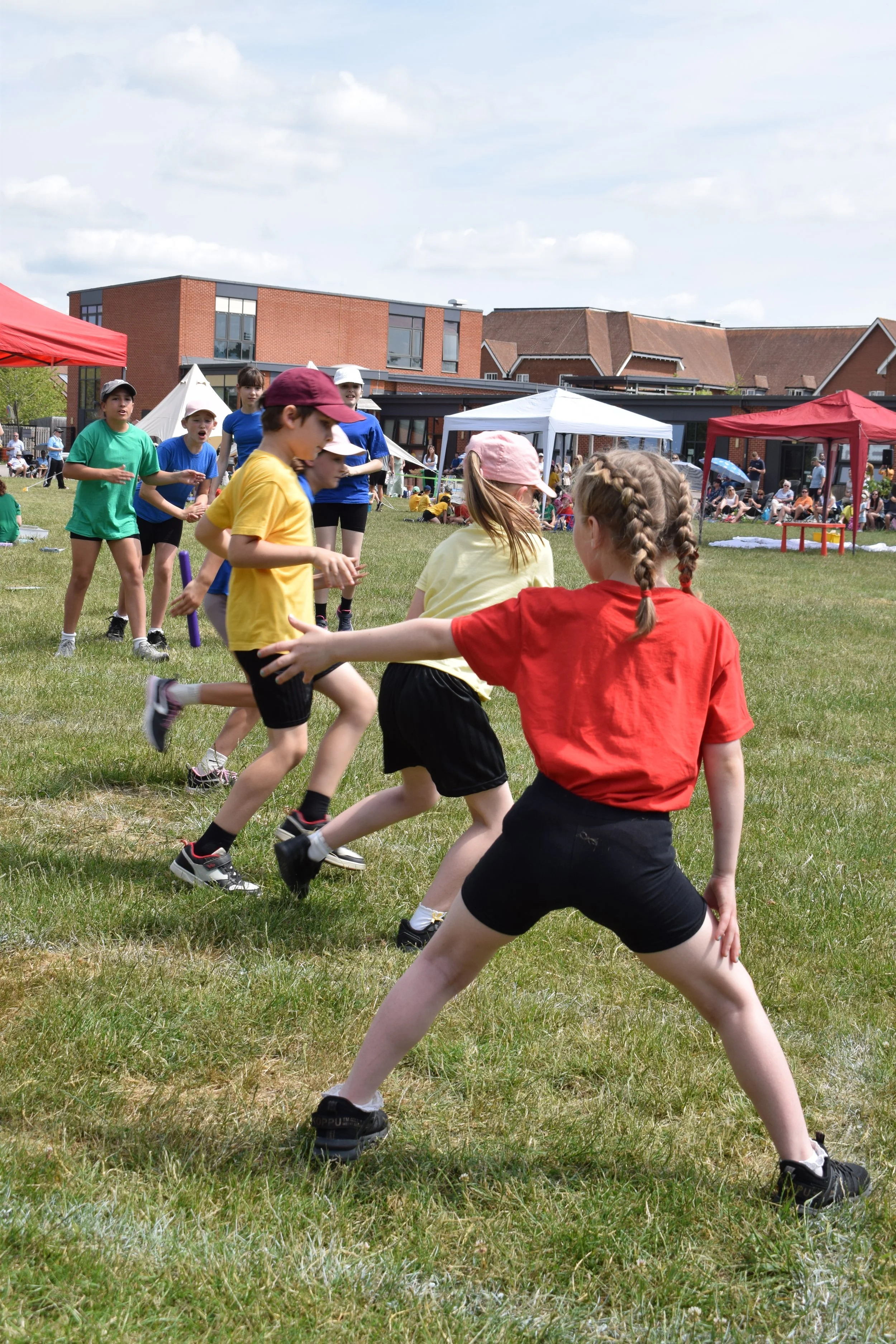 Children running and playing outdoors at a park or school field with tents and spectators in the background.