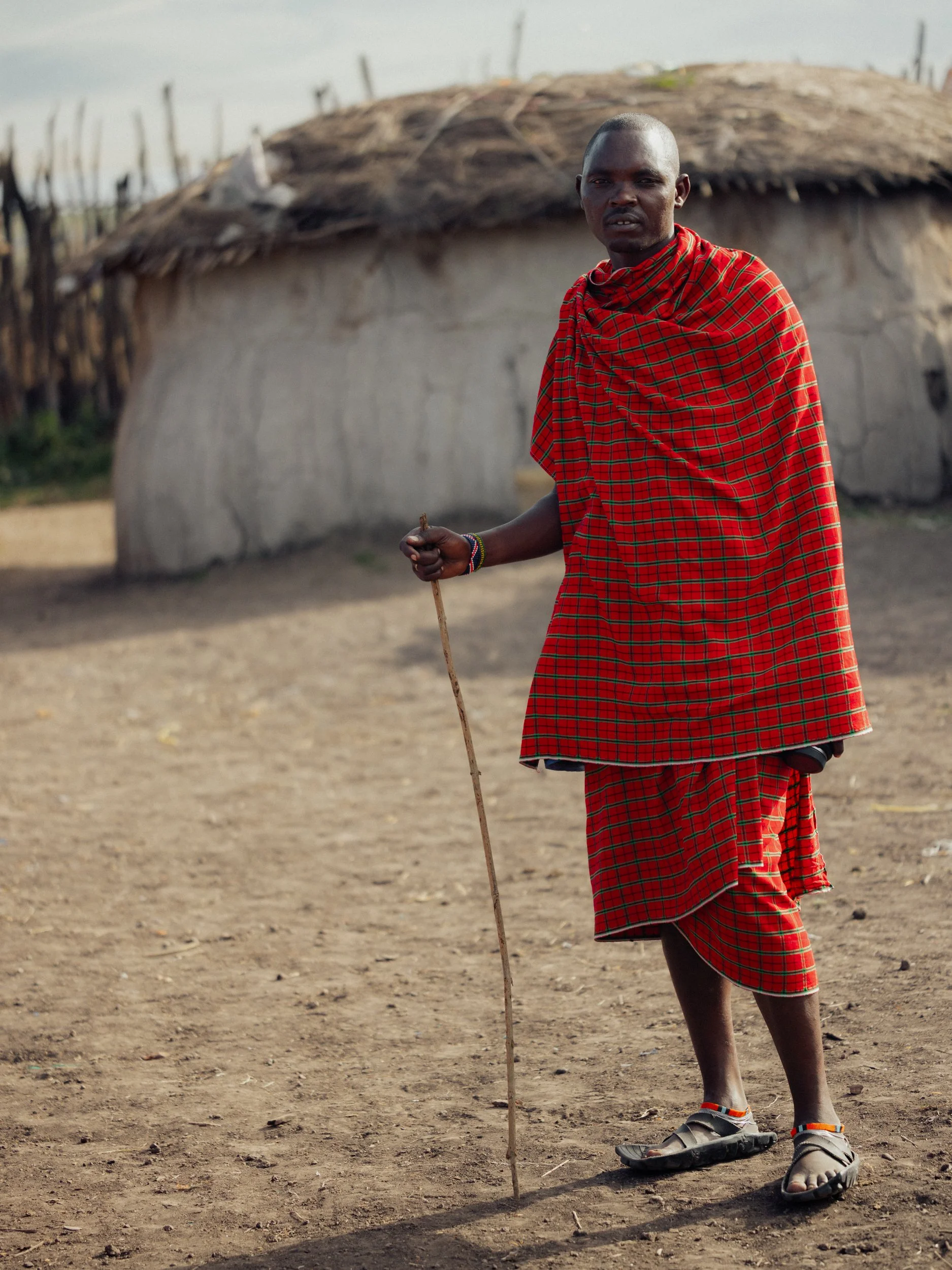 A Maasai man, wearing a red plaid shuka, standing outdoors on dry, dusty ground. He holds a stick in one hand and is wearing sandals. In the background, there is a small hut with a thatched roof and wooden fence. Photographed in Ngorongoro by Rob Ash