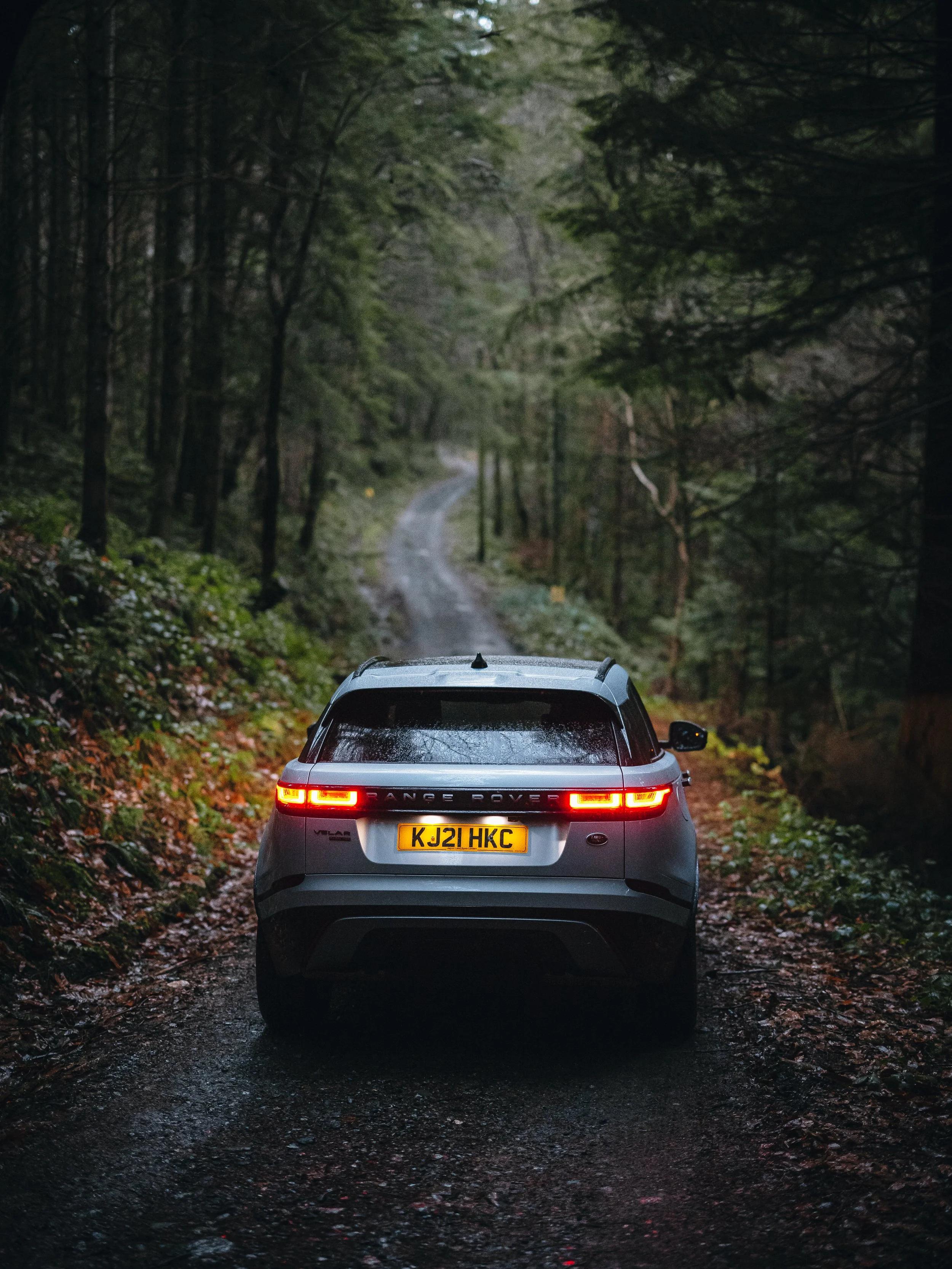 A silver Range Rover Velar driving along a narrow, muddy forest trail surrounded by dense trees and greenery, photographed by Rob Ash