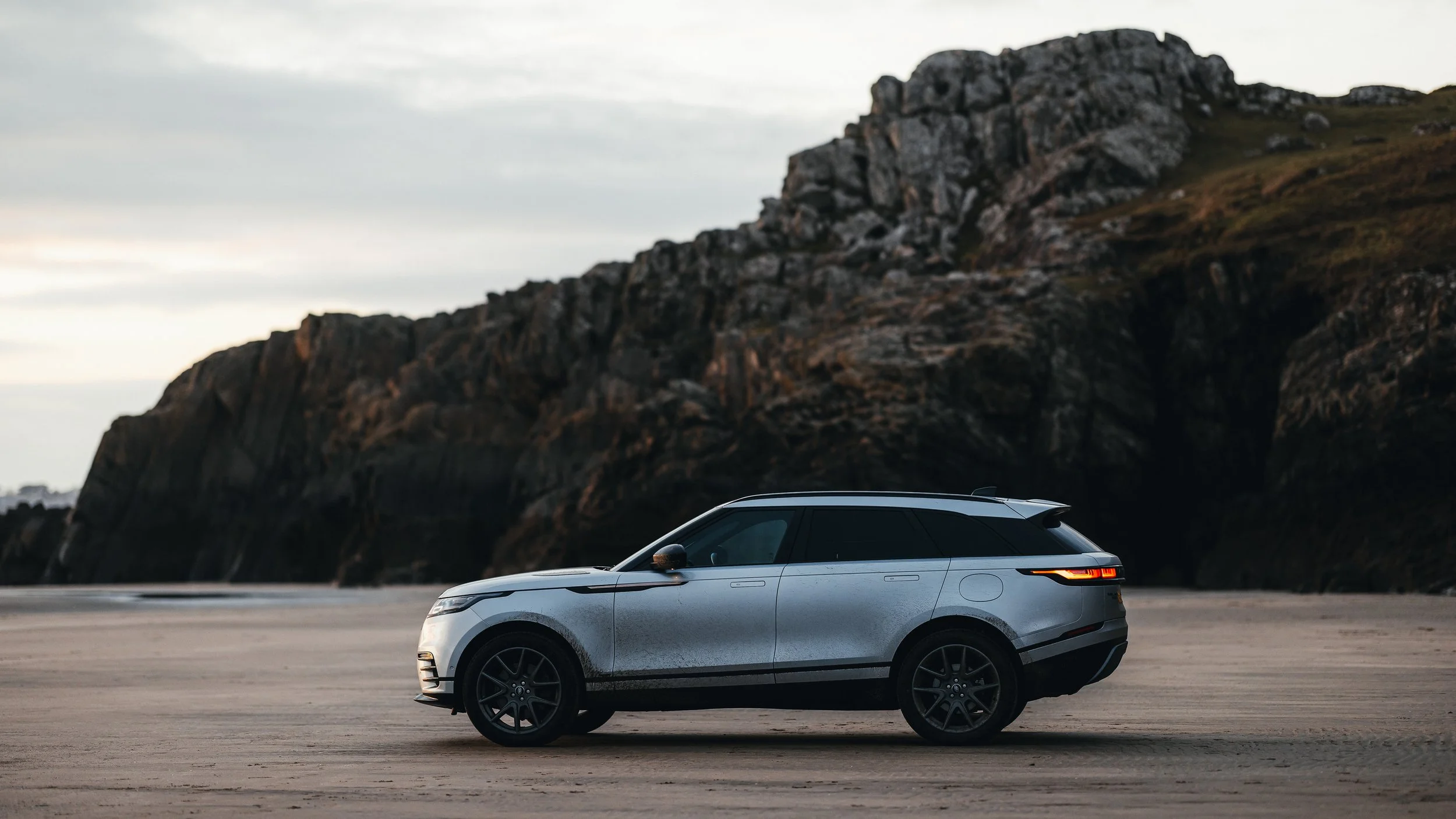 A silver Range Rover Velar parked on Black Sands beach in Wales, with rocky cliffs in the background during cloudy weather, photographed by Rob Ash
