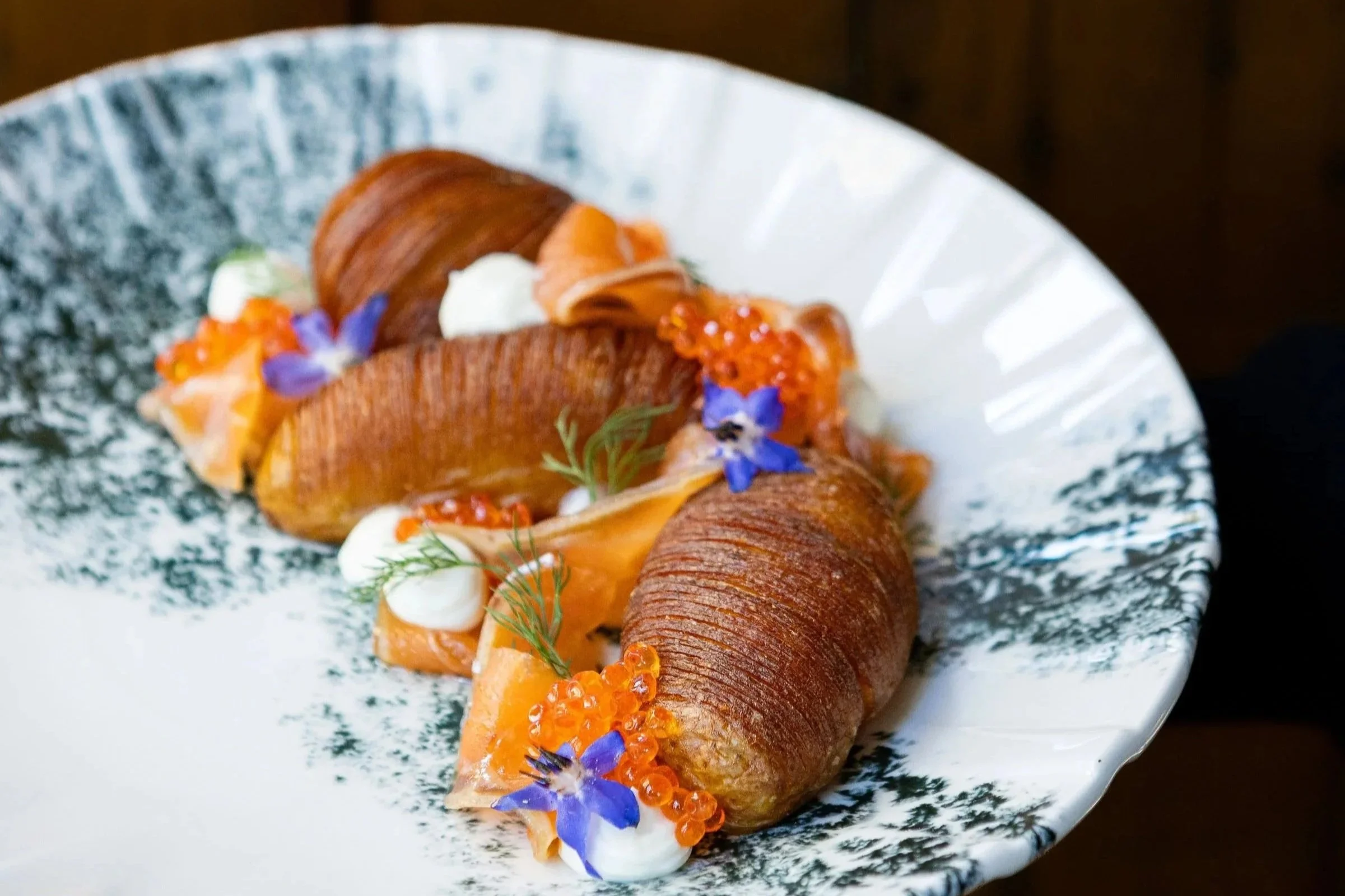 A gourmet dish on dark and light plate, including seafood, vegetables, bread, and garnishes, arranged on a dark surface.