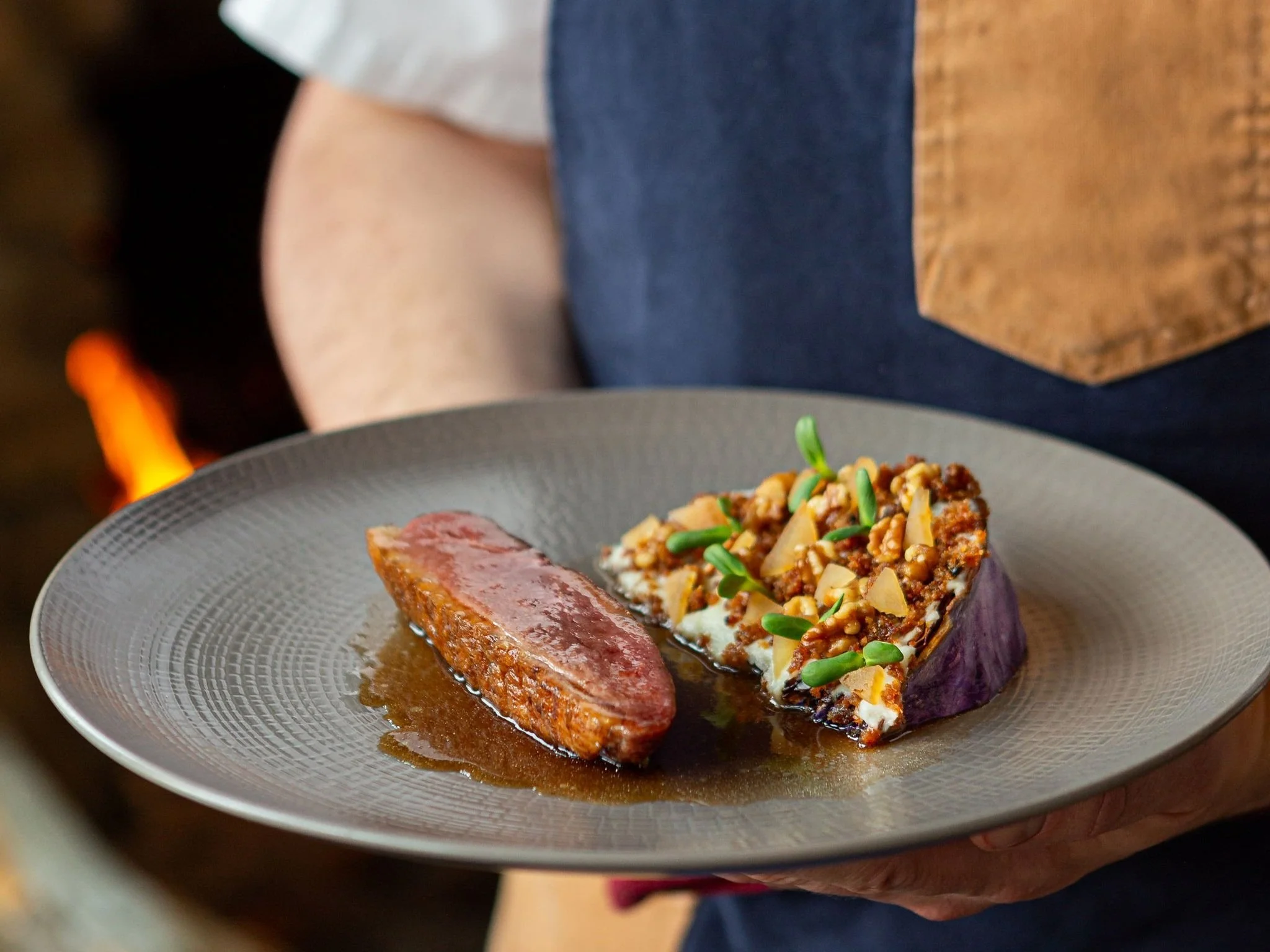 A person holding a gray plate with seared meat topped with sauce and a baked sweet potato half topped with chopped nuts, green sprouts, and sauce.