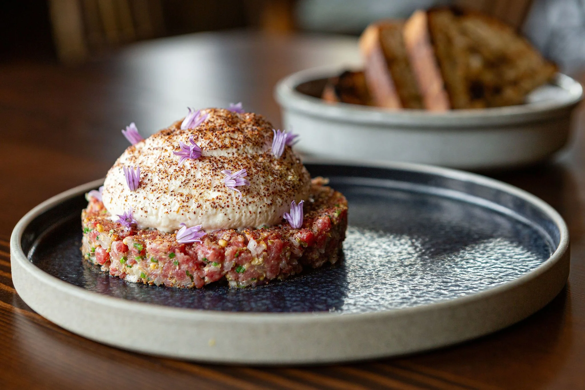 A plate of steak tartare topped with a dollop of creamy sauce and garnished with purple edible flowers, with a side of bread slices in a bowl in the background.