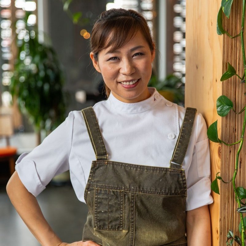 A smiling woman in a white chef's coat and brown apron standing indoors near a wooden wall with green leaves, with blurred background of green plants and interior decor.