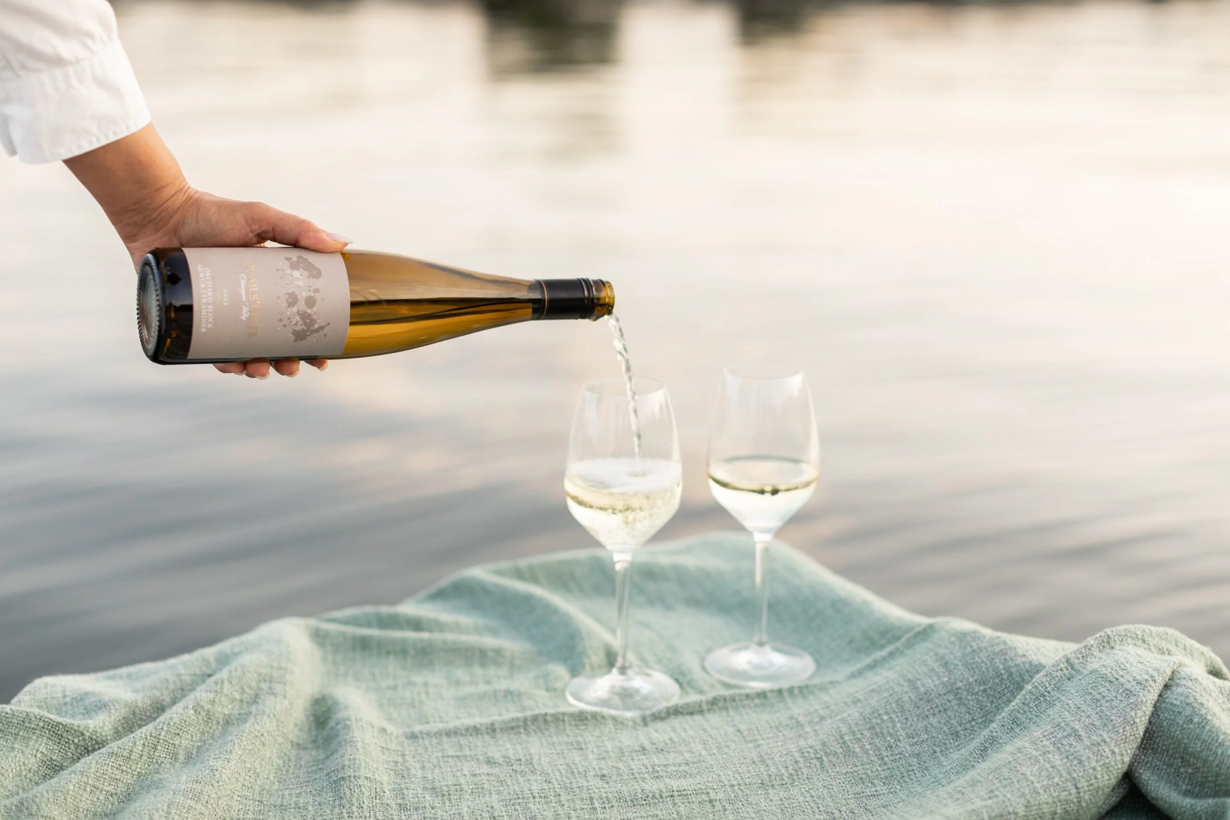 A person pouring white wine into a glass on a blanket by the water with another glass of wine upright nearby.