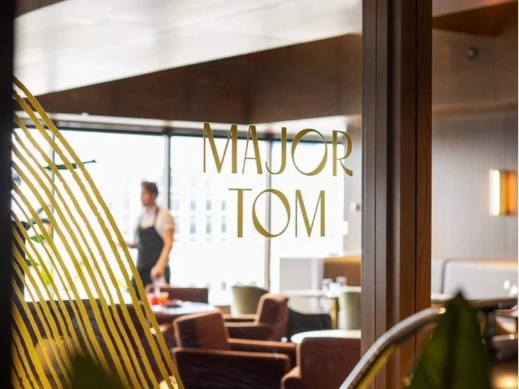 Interior of a restaurant named 'Major Tom,' with a waiter in the background, seating area with chairs, and a window showing city buildings.