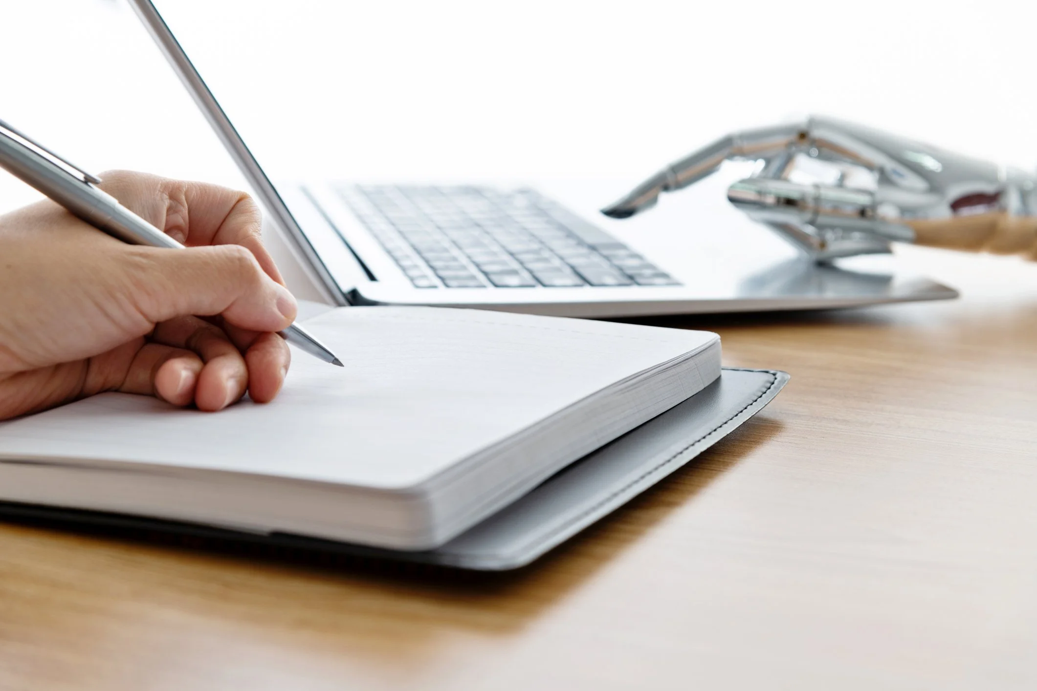 person writing with pen in a notebook next to robot hand typing on laptop to represent personal writing and AI writing
