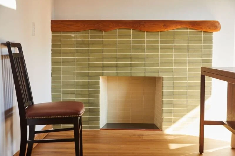 Interior view of a fireplace with green tiles, a wooden mantel, and surrounding furniture including a chair and a table.