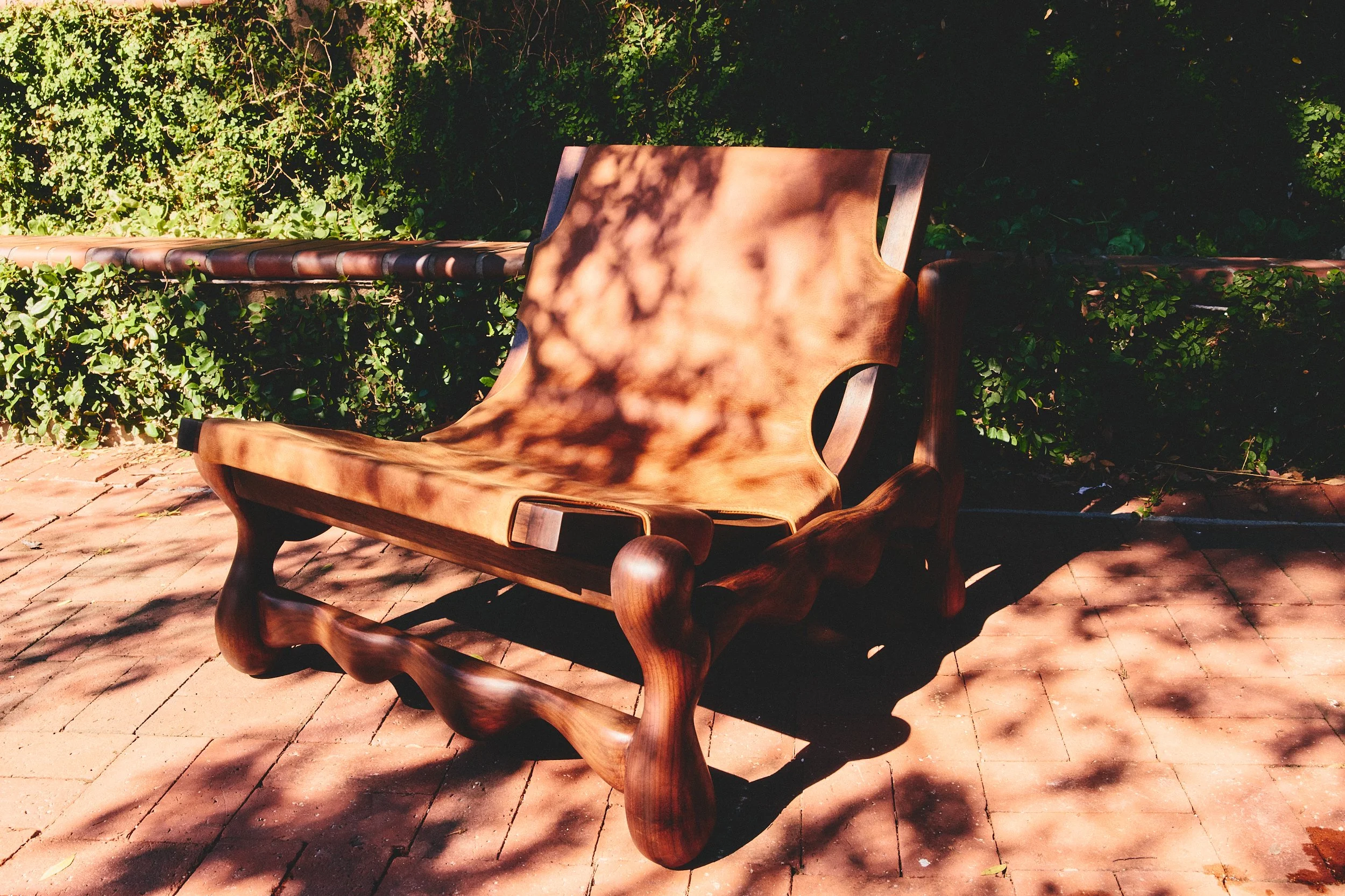 Wooden bench with a leather cushion, shaded by tree branches, on a brick patio with green bushes in the background.