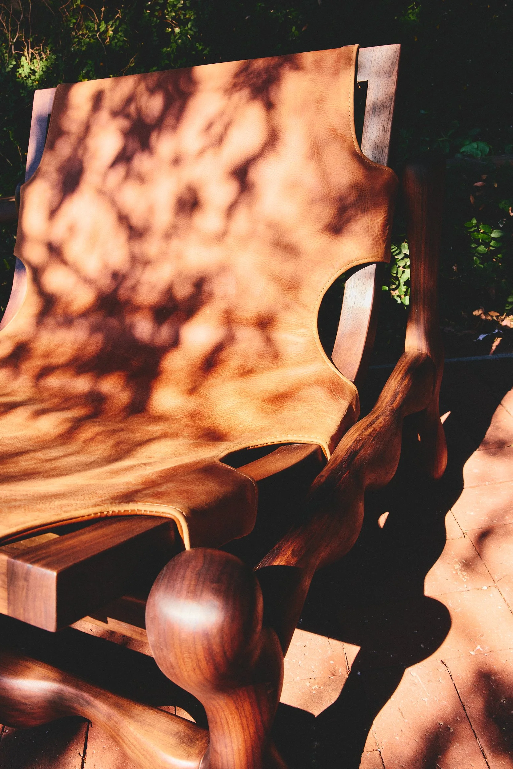 A wooden bench with a leather or faux leather seat in a garden setting, with dappled sunlight and shadows from nearby trees.