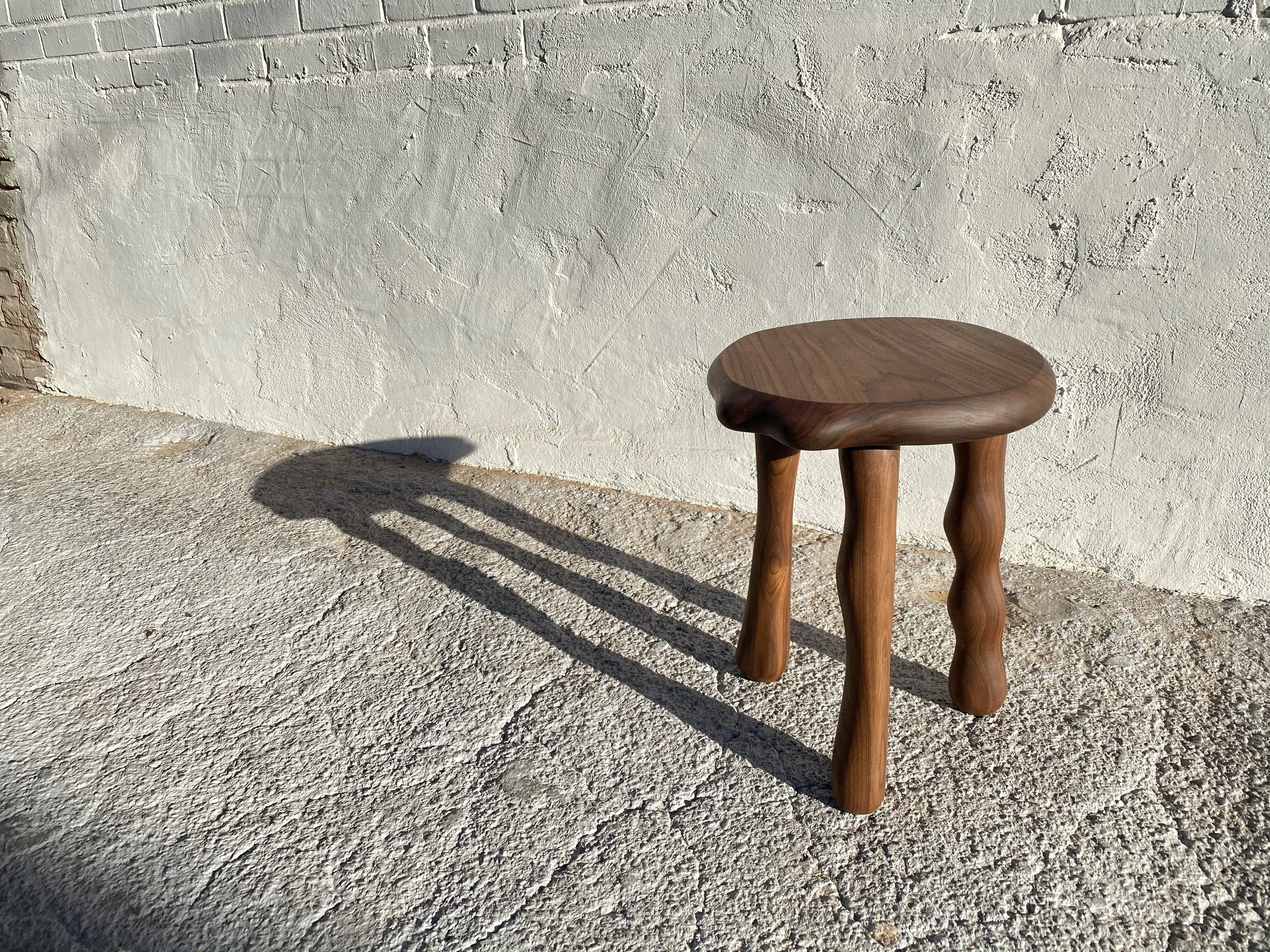 A wooden stool with three curved legs, placed outdoors on a textured concrete surface, casts a shadow on the ground. Behind, there is a white textured wall with visible cracks and patches.