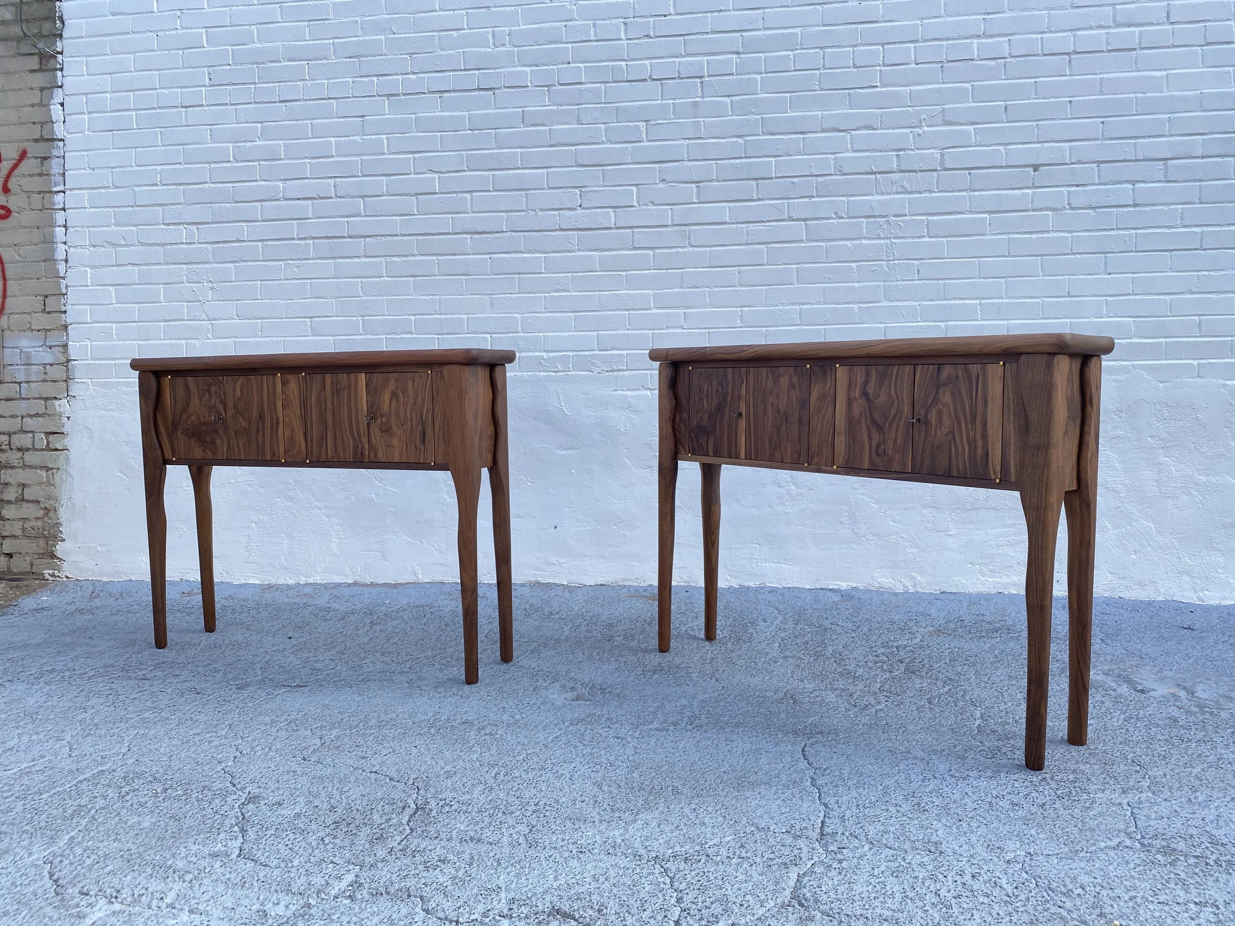 Two wooden side tables are placed against a white brick wall, with parts of a brick wall and graffiti visible on the left side. The ground is concrete with visible cracks.