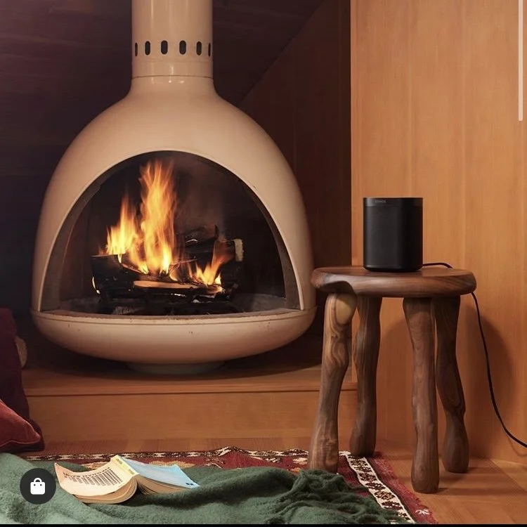 A cozy living room corner with a modern, rounded beige fireplace burning fire, next to a small wooden side table holding a black speaker, and a rug with an open book and green blanket in the foreground.