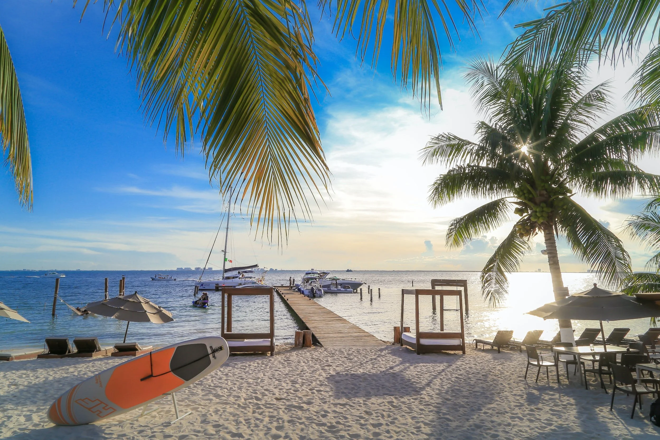 A tropical beach scene with palm trees, a wooden pier extending into the water, sailboats and motorboats, beach chairs, umbrellas, and a paddleboard on the sandy shore, with the sun setting or rising over the ocean.
