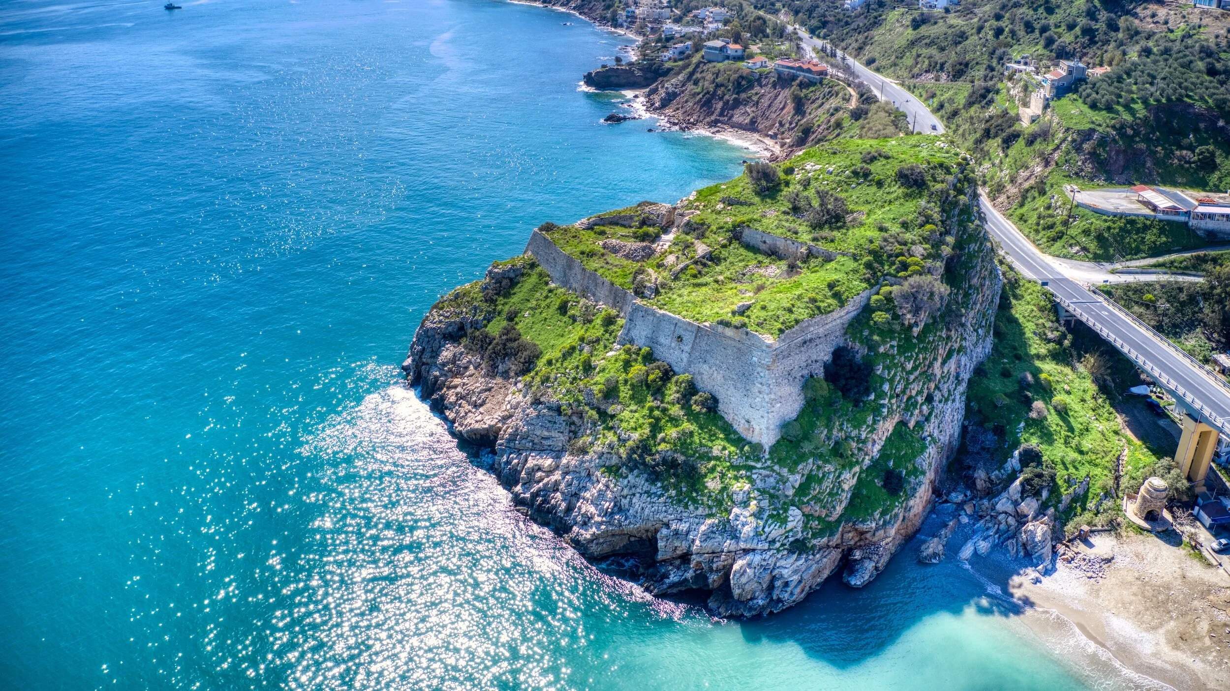 Aerial view of a historic stone fortress built on a large rocky island off the coast, surrounded by bright blue ocean waters, with a winding road along the hillside.