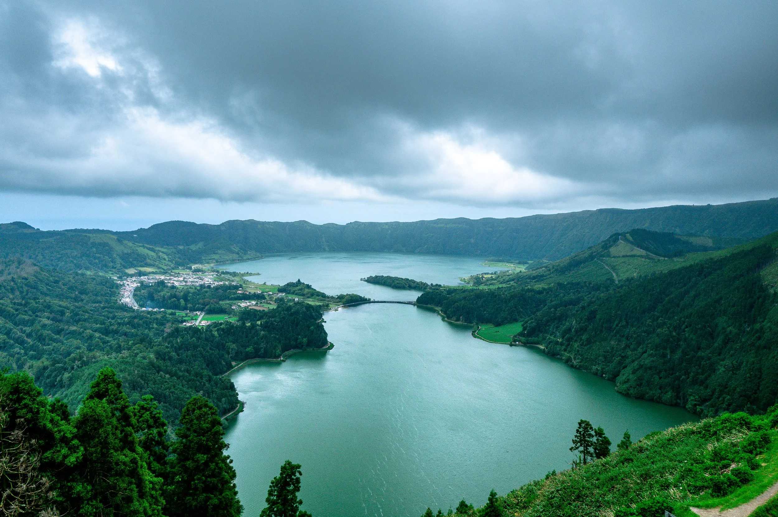 A view of a large lake surrounded by lush green forests and hills under a cloudy sky.