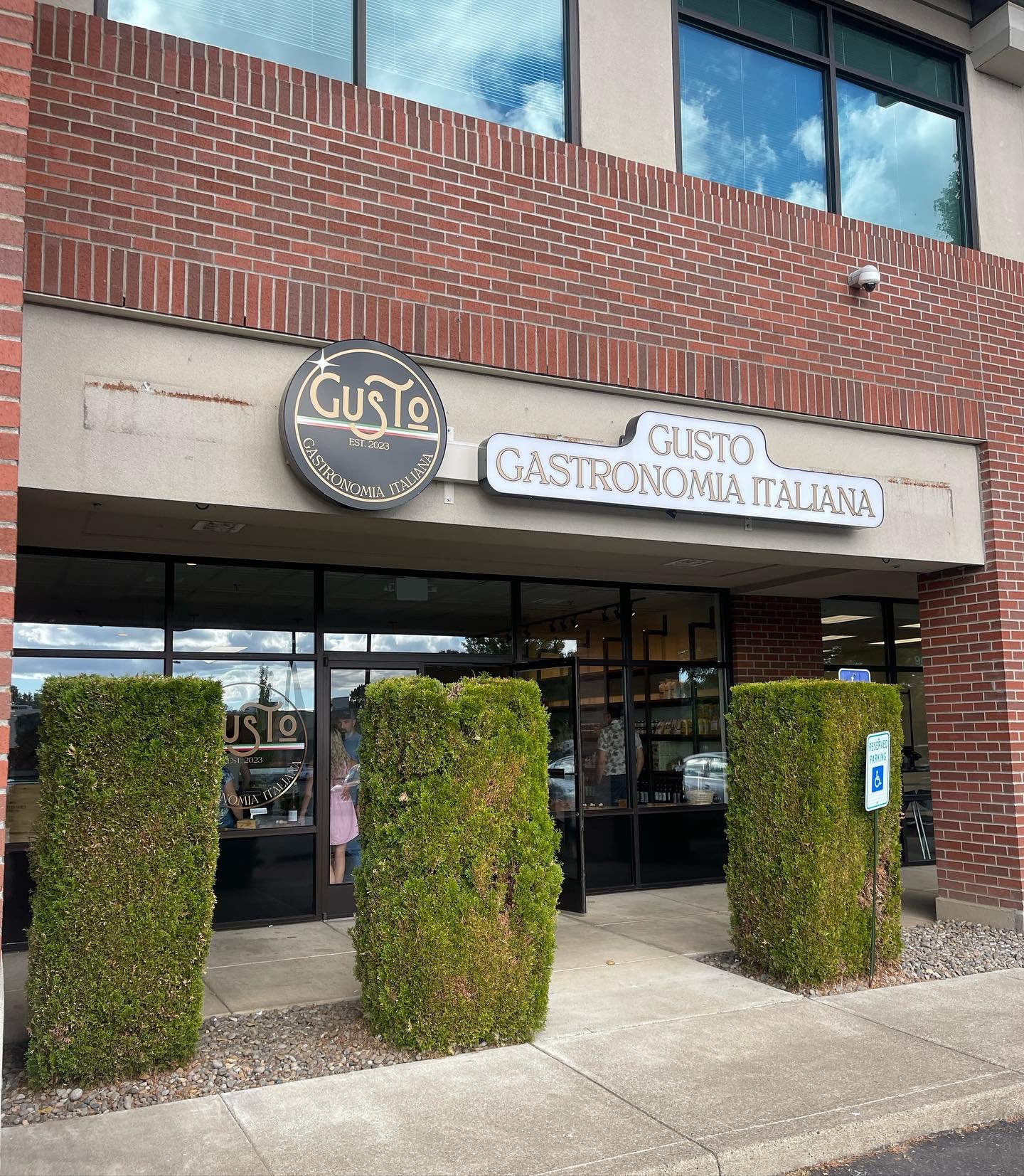 The storefront of Gusto, an Italian restaurant, with the sign 'Gusto Gastronomia Italiana'. There are three trimmed bushes outside, a handicap parking sign, and large windows showing the interior and people inside.