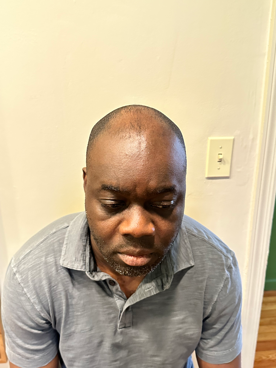 A middle-aged man with dark skin and a short beard looking downward, wearing a gray polo shirt, standing indoors against a pale wall and floor.