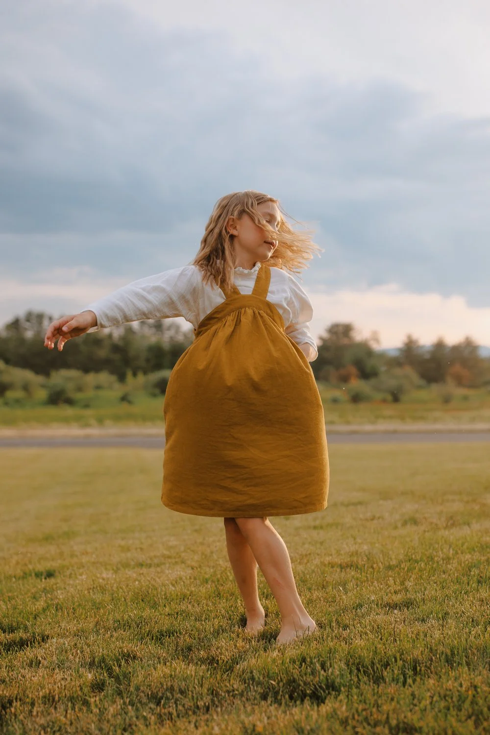 A young girl in a brown dress spinning in an open grassy field under a cloudy sky.