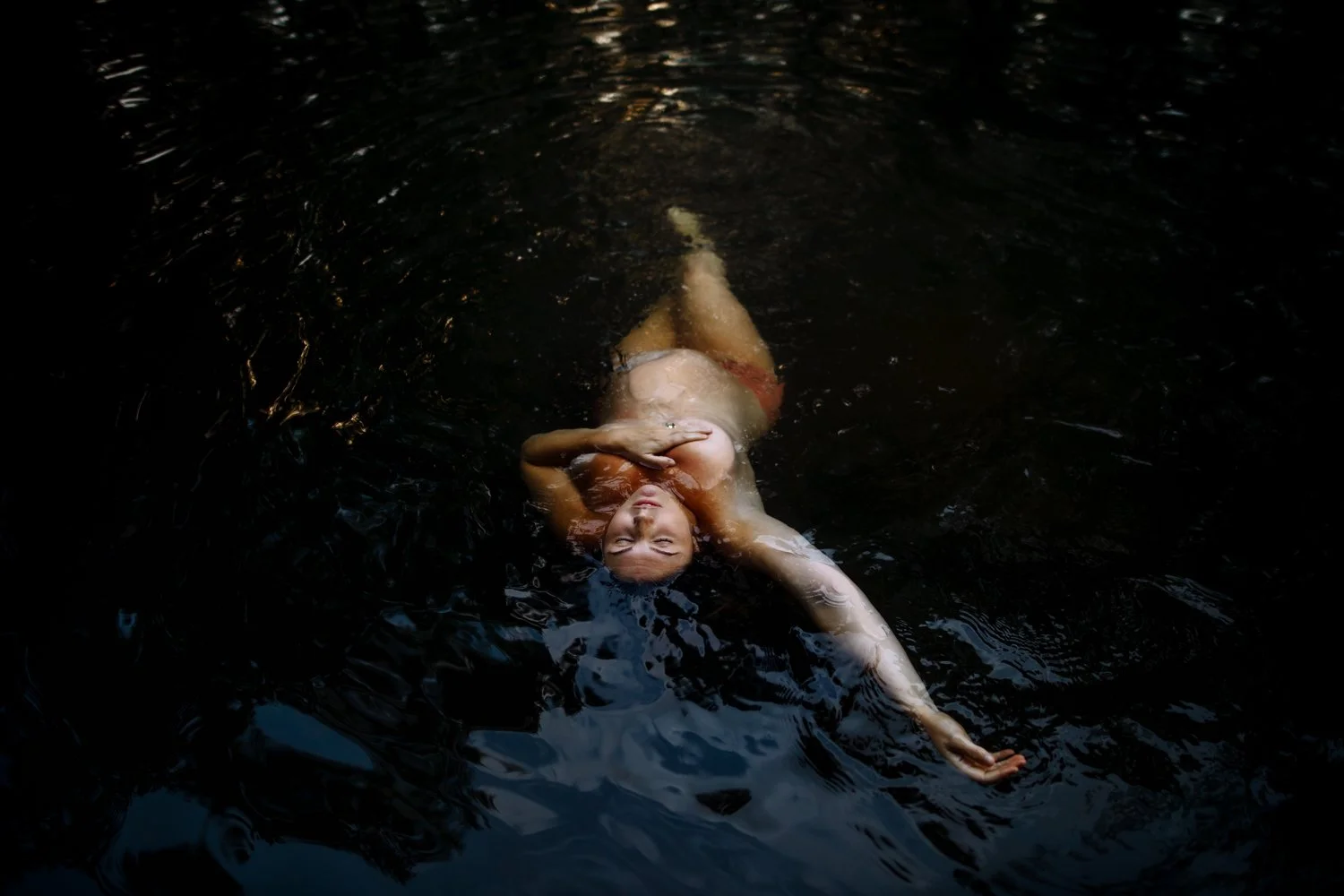 Pregnant woman floating in dark water with her arms raised overhead