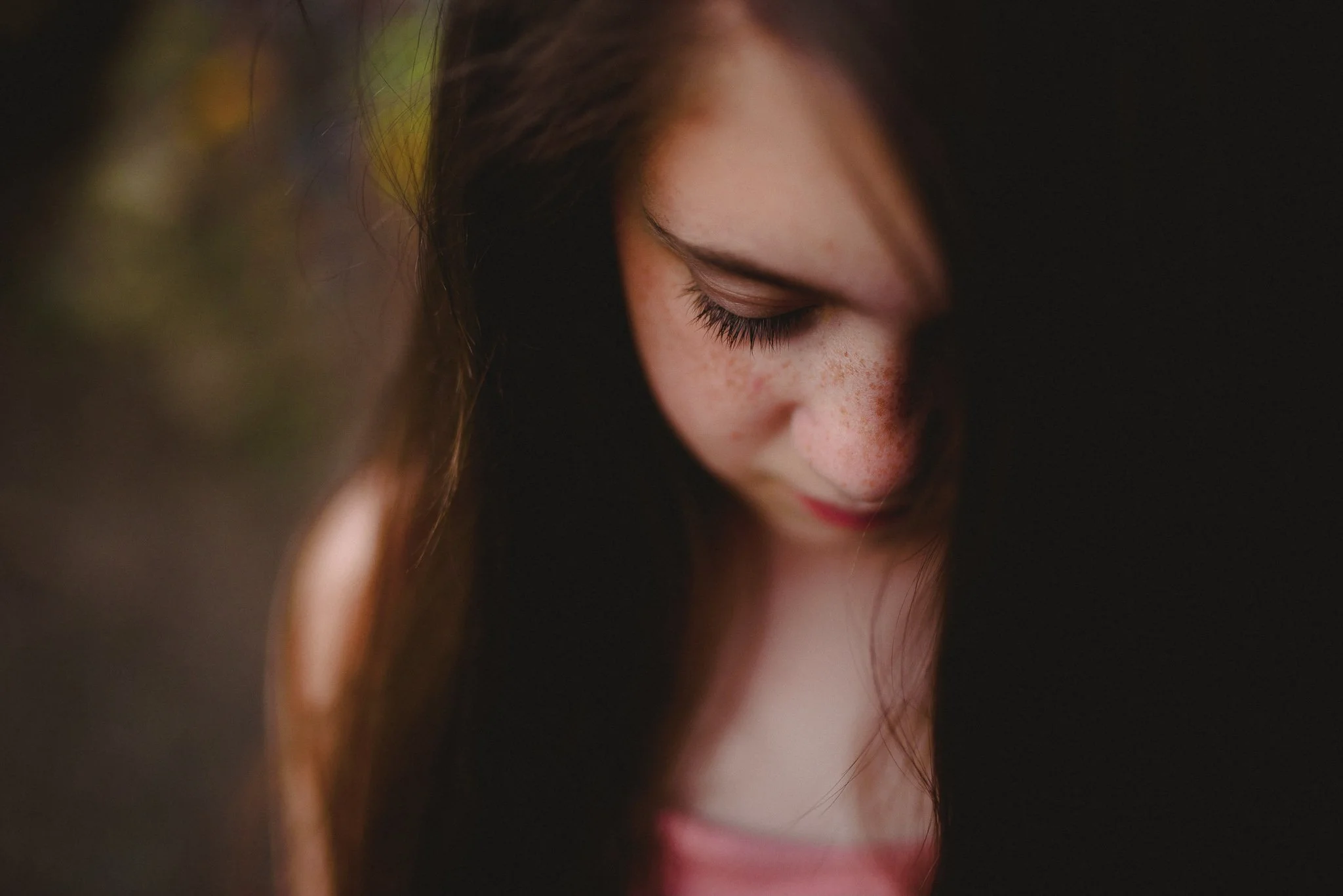 Close-up of a young girl with freckles, looking down with her eyes closed, soft lighting, dark background.