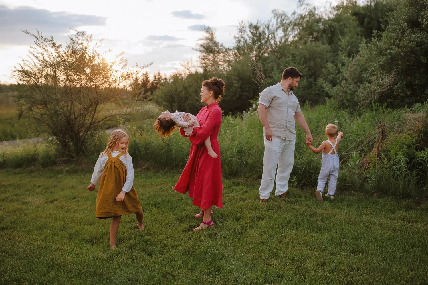 Family of five walking through a grassy field at golden hour, with mother swinging her toddler and father holding their son's hand