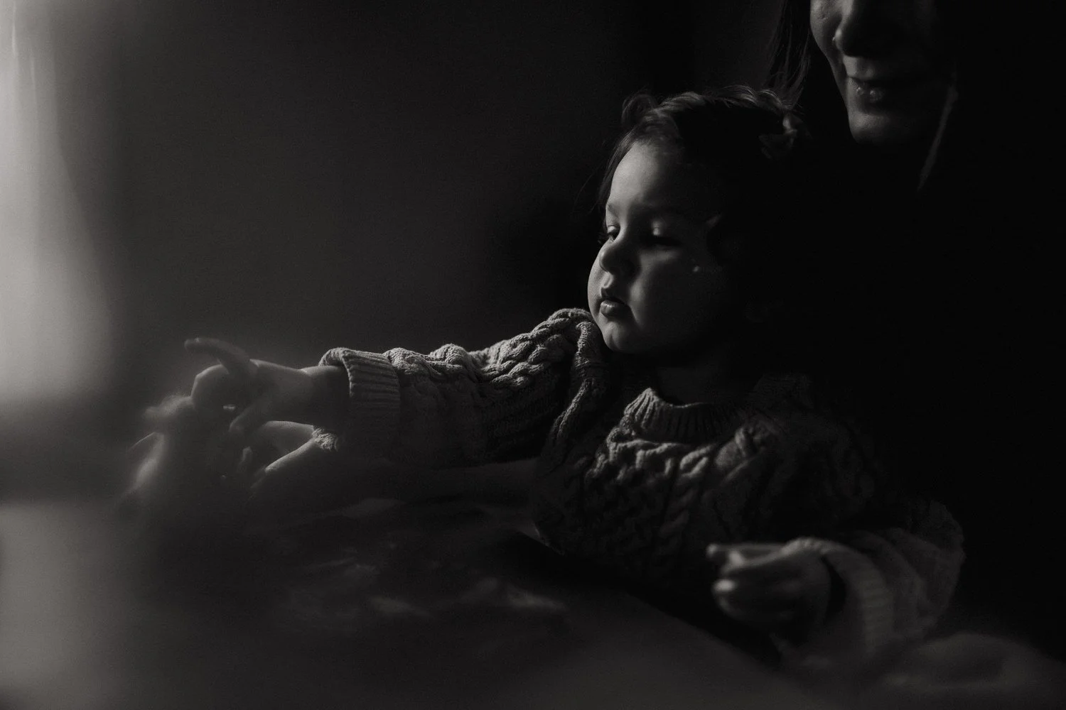 Black and white portrait of a toddler reaching forward with her mother softly lit behind her
