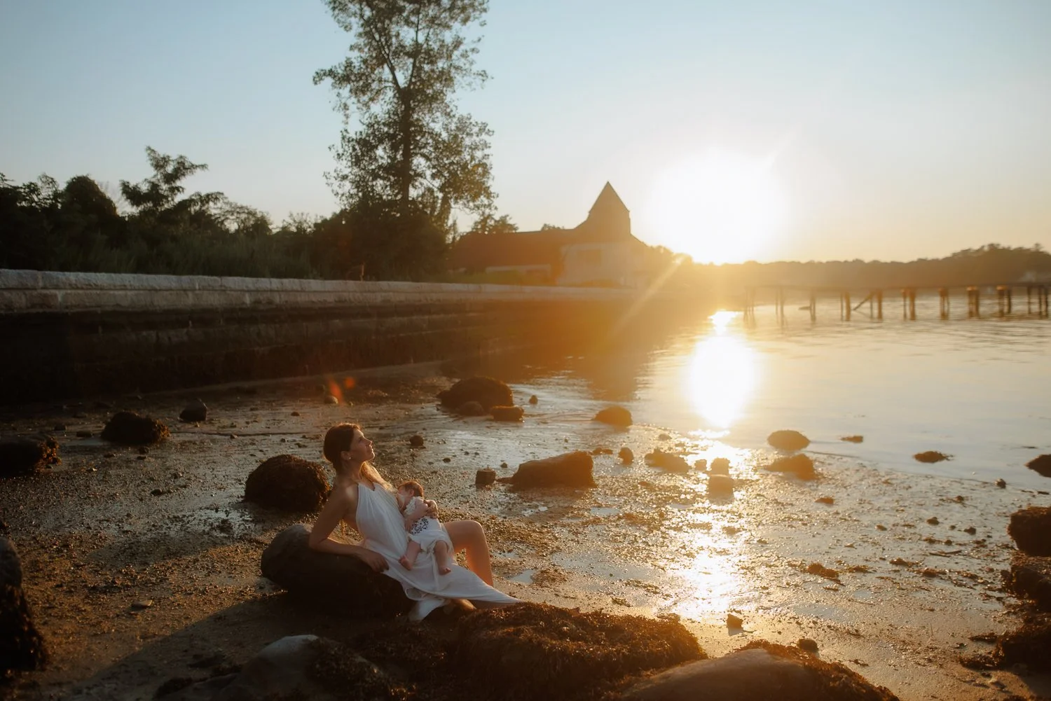 Mother sitting on rocks at the Bay holding her baby at golden hour, waterfront session with a baby photographer in New York