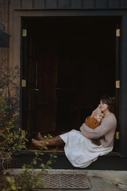 Mother sitting in an open doorway holding her toddler close, both wrapped in a cozy embrace.