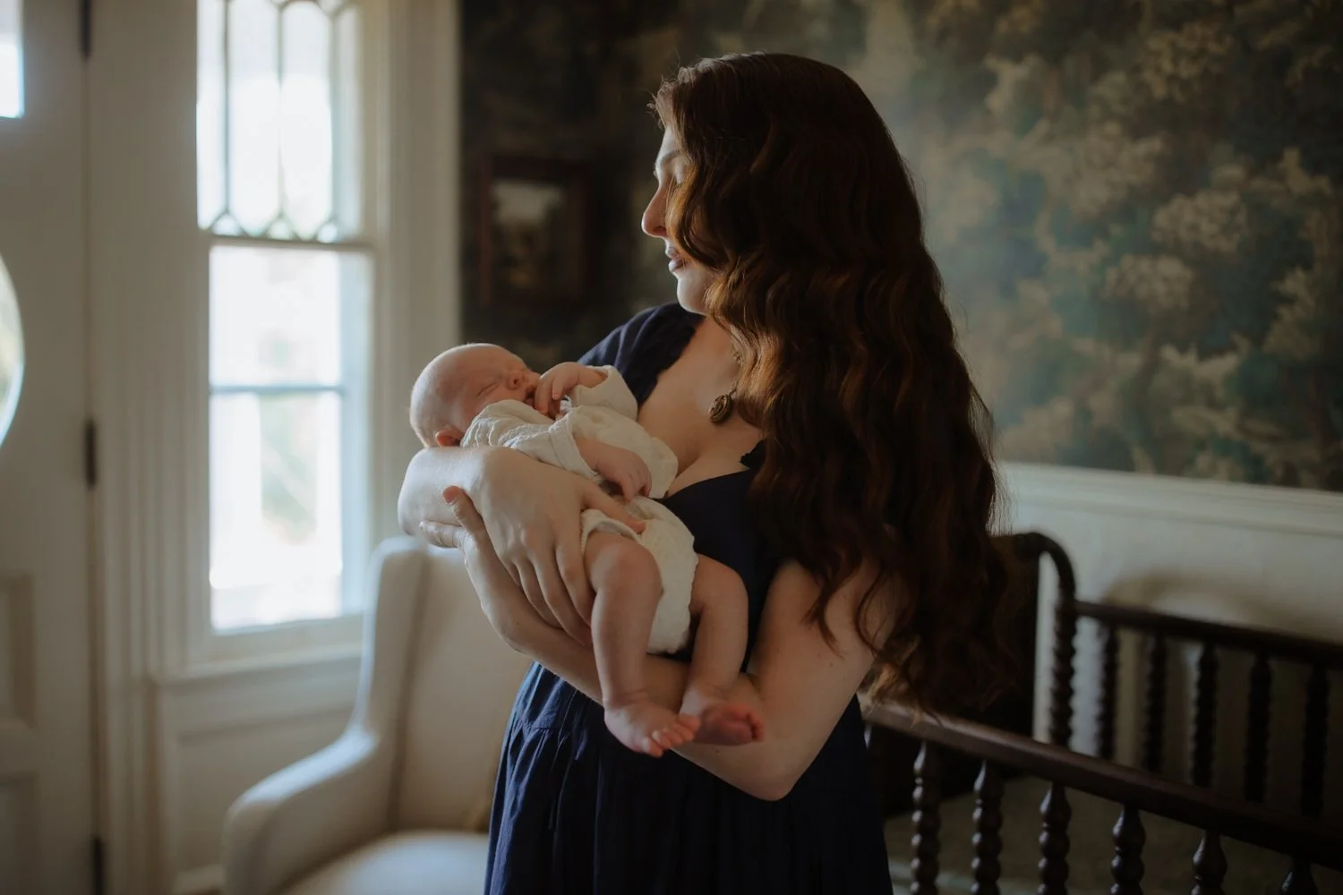 Mother gazing out a nursery window while holding her newborn, New York newborn photography session